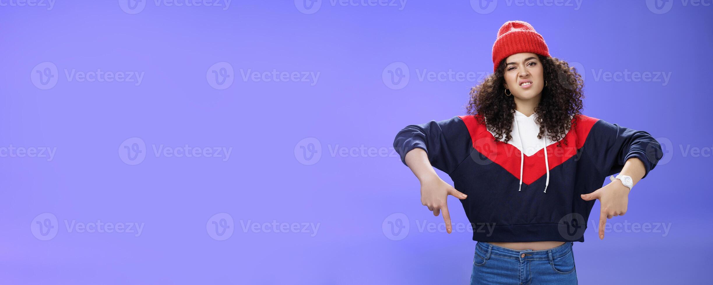 Snobbish and displeased stylish and cool street girl with curly hair in beanie and sweatshirt pointing down grimacing as feeling unsatisfied and disappointed posing against blue background photo