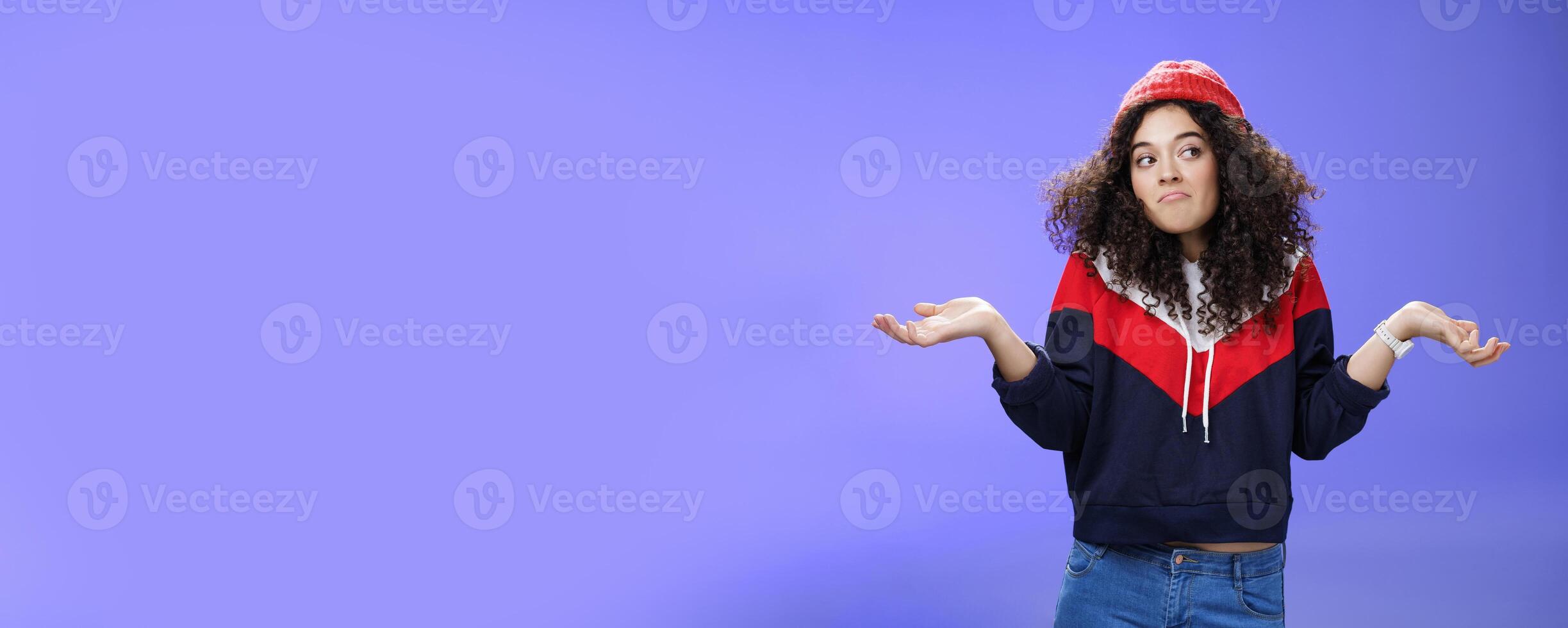 Not getting clue. Cute silly woman with curly hair shrugging with pursed lips and unbothered expression holding hands sideways looking right being unaware and confused, uncertain what answer photo