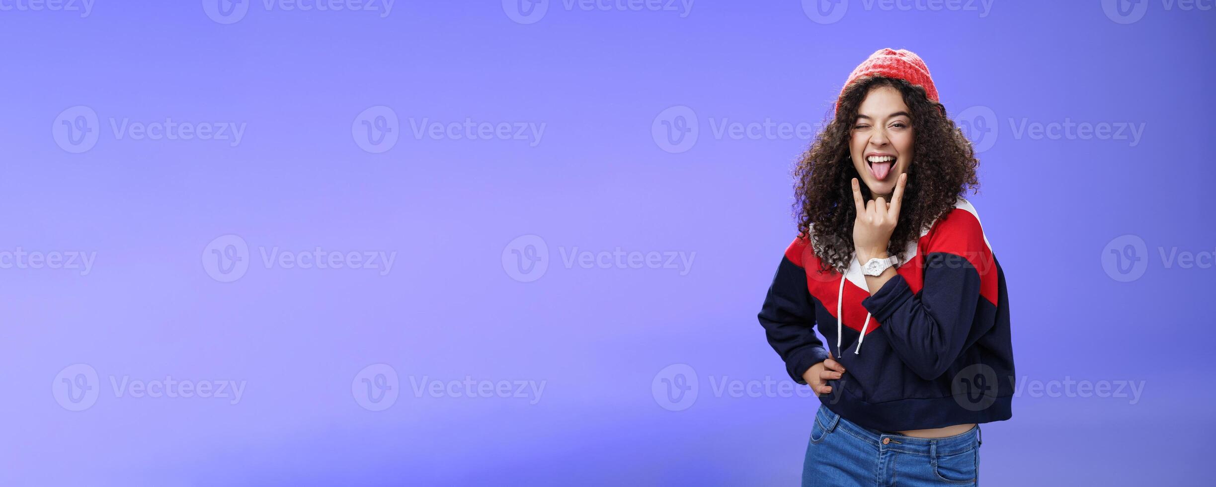 Stylish and cool energized swag girl with curly hairstyle in warm beanie showing rock-n-roll gesture near mouth and sticking out tongue smiling having fun, partying and enjoying life over blue wall photo