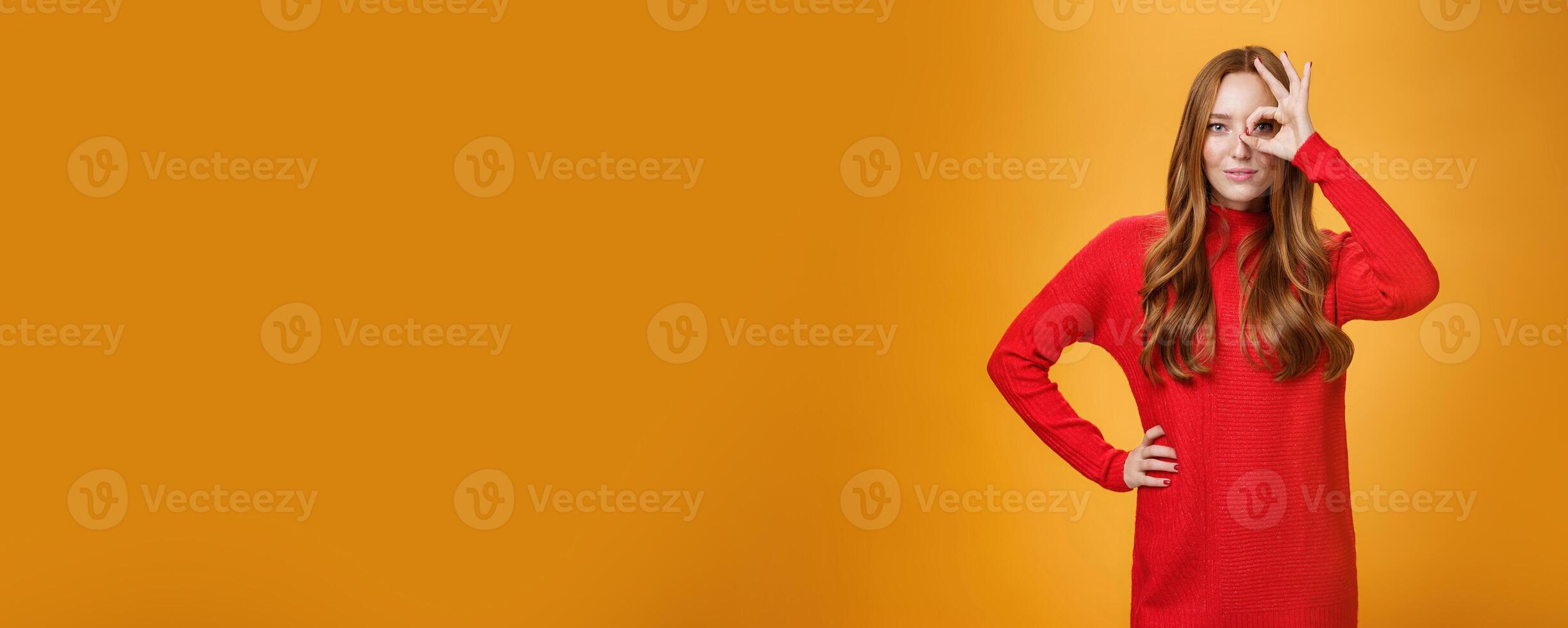 I got you on aim. Self-assured expressive and confident attractive redhead woman in red warm dress showing okay or zero sign on eye and peeking through as smiling satisfied over orange wall photo