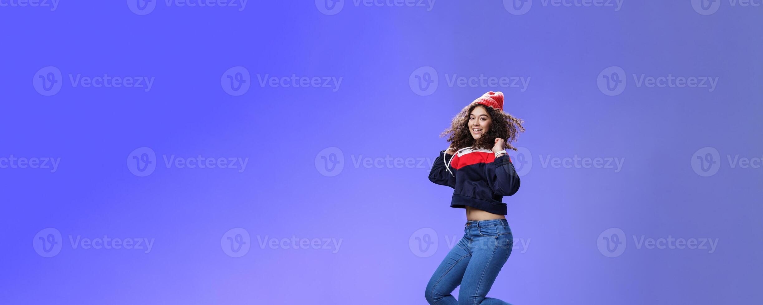 Studio shot of cute girl with curly hair in beanie sweatshirt and sneakers jumping playful and carefree over blue background, having fun enjoying cool weather smiling broadly as flying in air photo