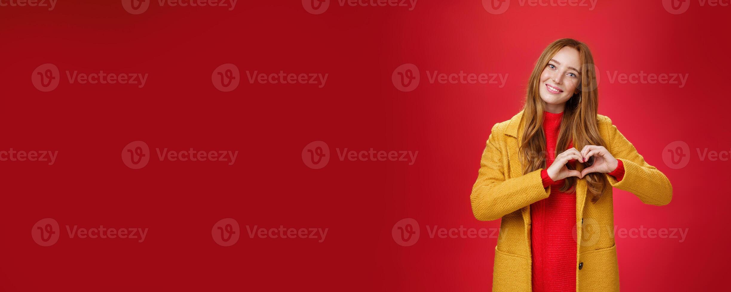 Love, romance and fall concept. Portrait of charming tender and gentle young redhead woman in yellow coat showing heart gesture making confession in sympathy, smiling cute at camera over red wall photo