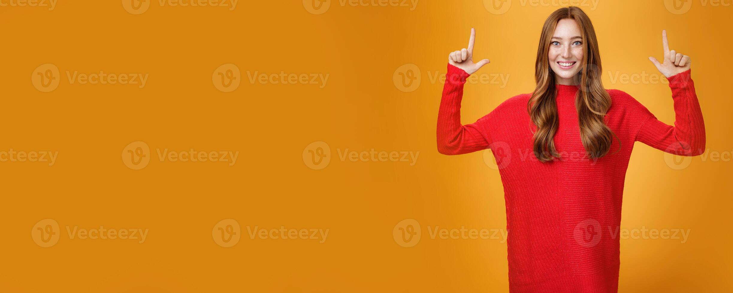 Enthusiastic and excited energized redhead female in red warm sweater pointing up with rised hands and smiling broadly thrilled with awesome promotion, feeling astonished against orange background photo