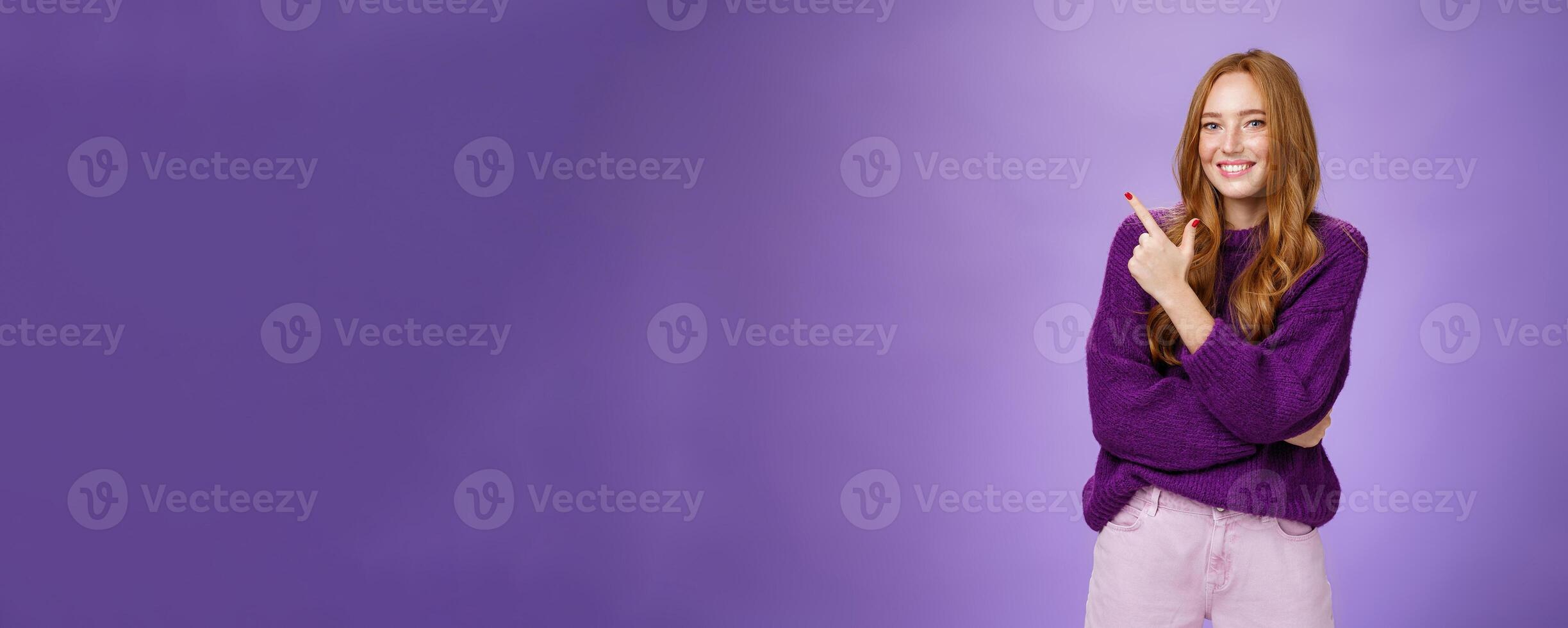 Charming friendly-looking bright redhead woman with freckles and makeup in purple warm sweater pointing at upper left corner and smiling delighted and cute at camera as showing cool place hang out photo