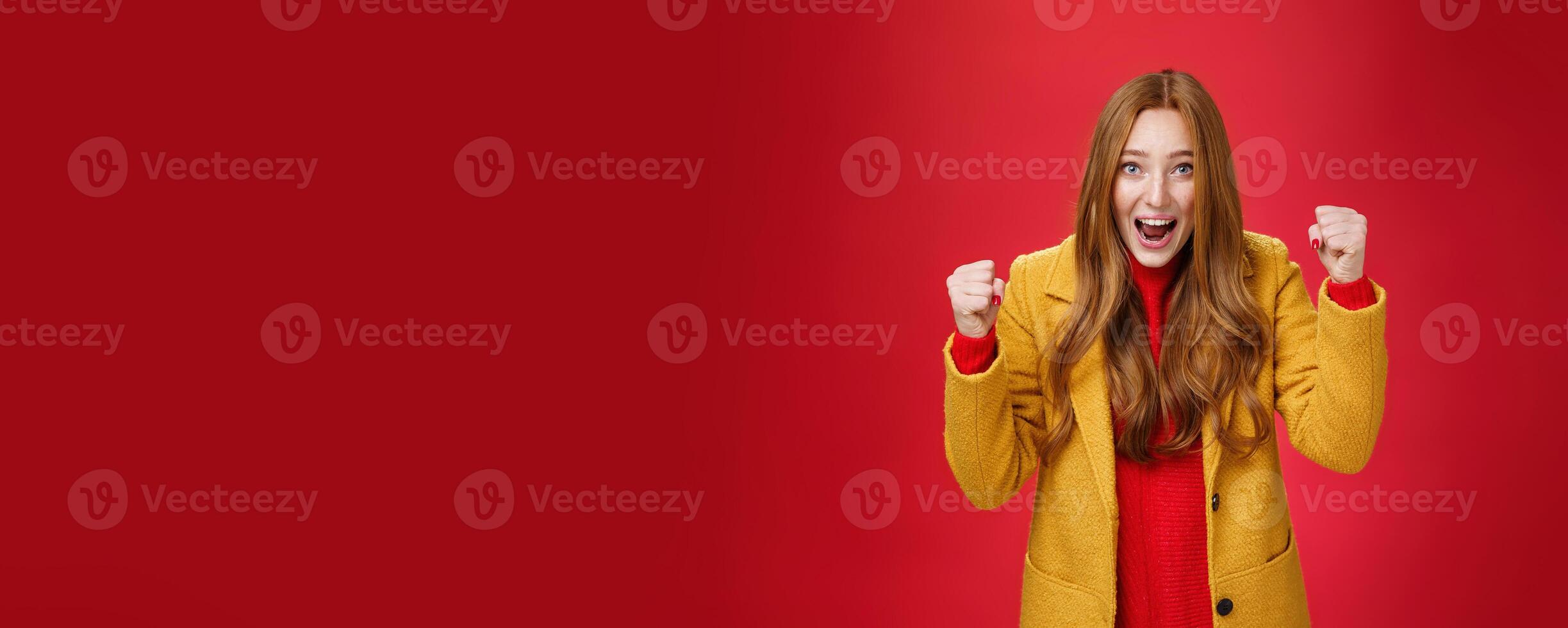Studio shto of cheerful excited good-looking ginger girl in yellow coat raising clenched fists in joy and happiness, triumphing yelling yes from success and triumph, celebrating win over red wall photo