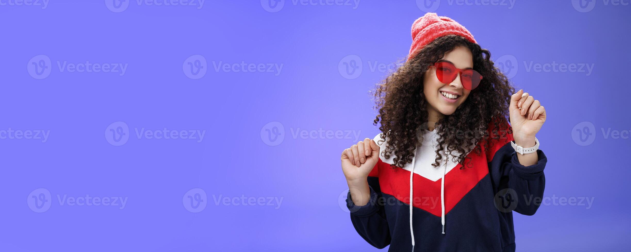 Stylish and feminine girlfriend dancing having fun feeling upbeat as liking weather feeling warm and cozy in stylish red beanie and sunglasses dancing with raised hands and broad smile over blue wall photo