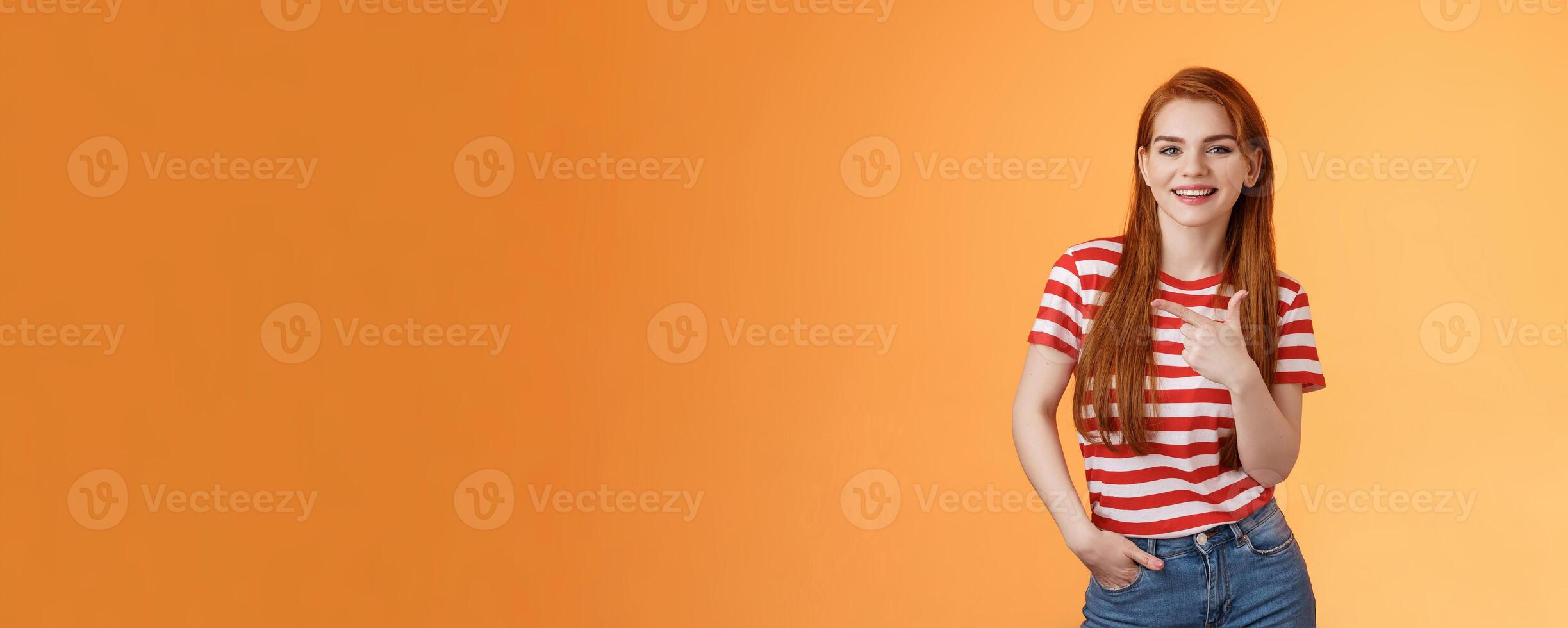 Carefree modern 20s female student chatting friends discuss left copy space, pointing left relaxed casual pose, smiling camera joyfully, introduce advertisement product, orange background photo