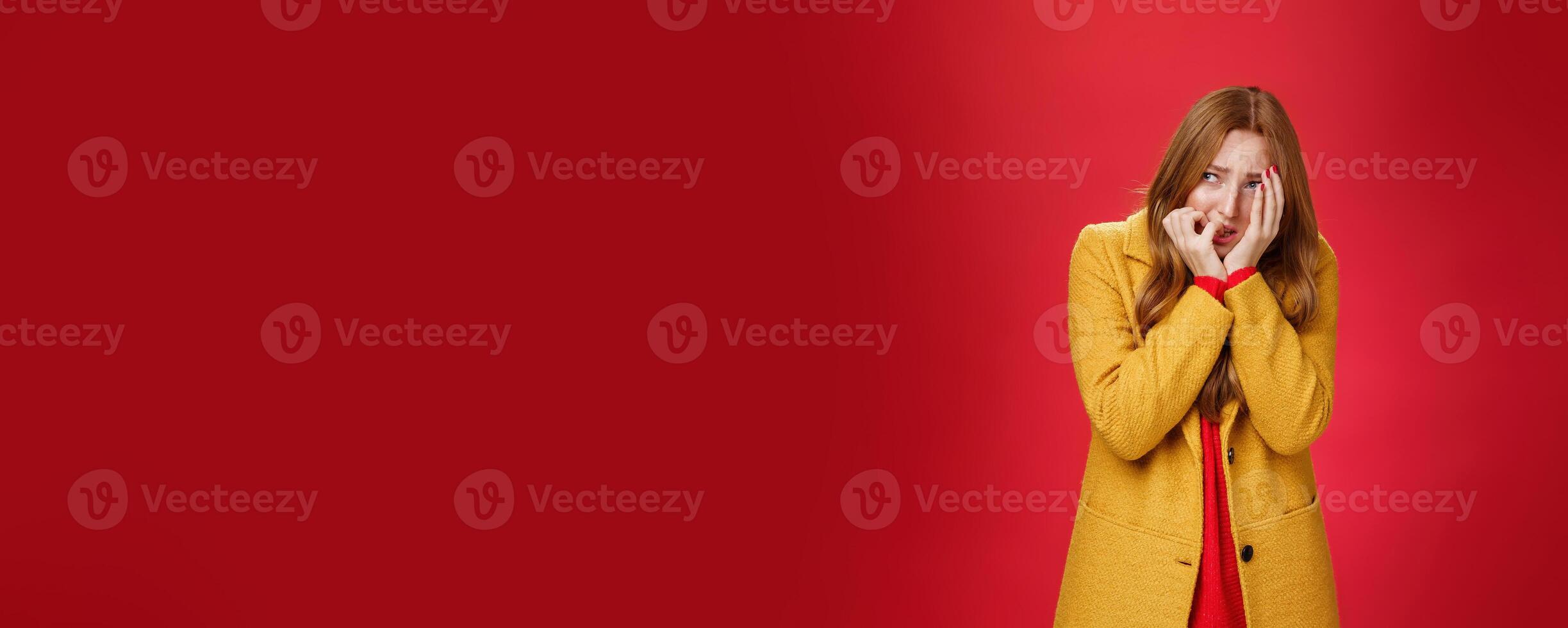 Portrait of shocked and scared insecure woman in yellow coat covering face in horror and fear biting nails squinting and looking intense left as frightened being victim standing in shook, trembling photo