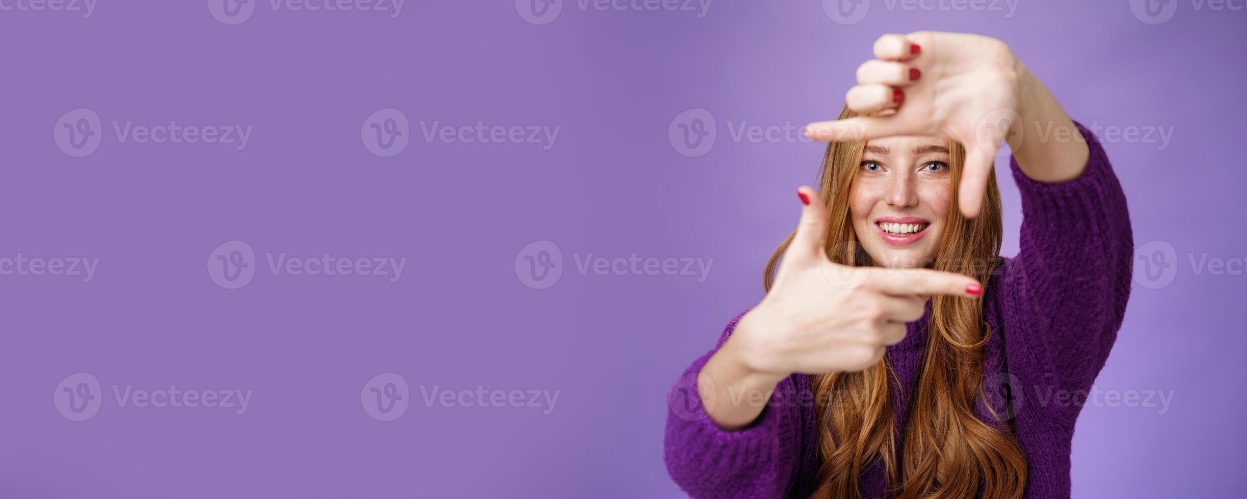 Imagine if. Portrait of hopeful and carefree creative charming redhead 20s woman showing frame with pulled hands and smiling through it as picturing bright and happy future over purple background photo