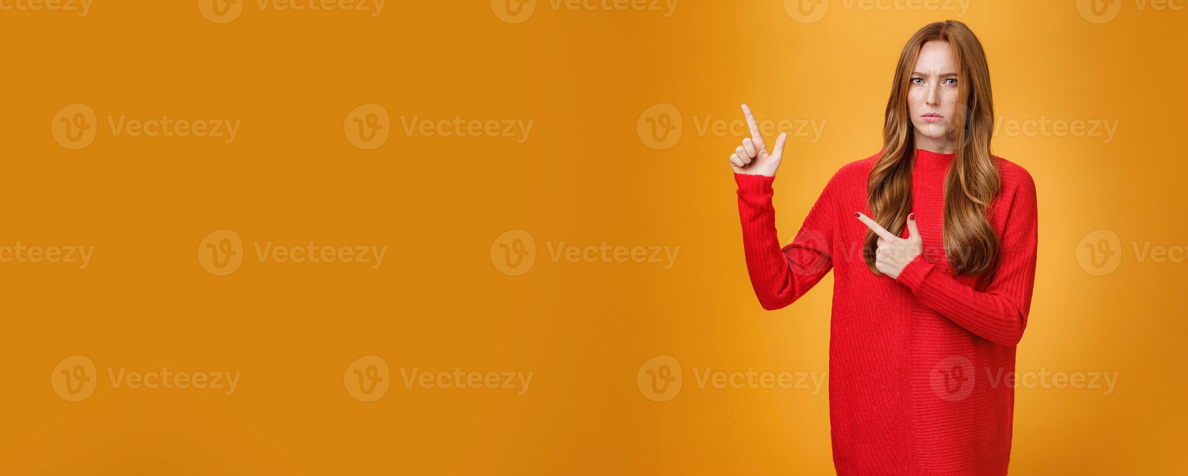 Intense suspicious and doubtful european ginger girl in warm sweater frowning hesitant and confused pointing at upper left corner asking question, looking with disbelief over orange background photo