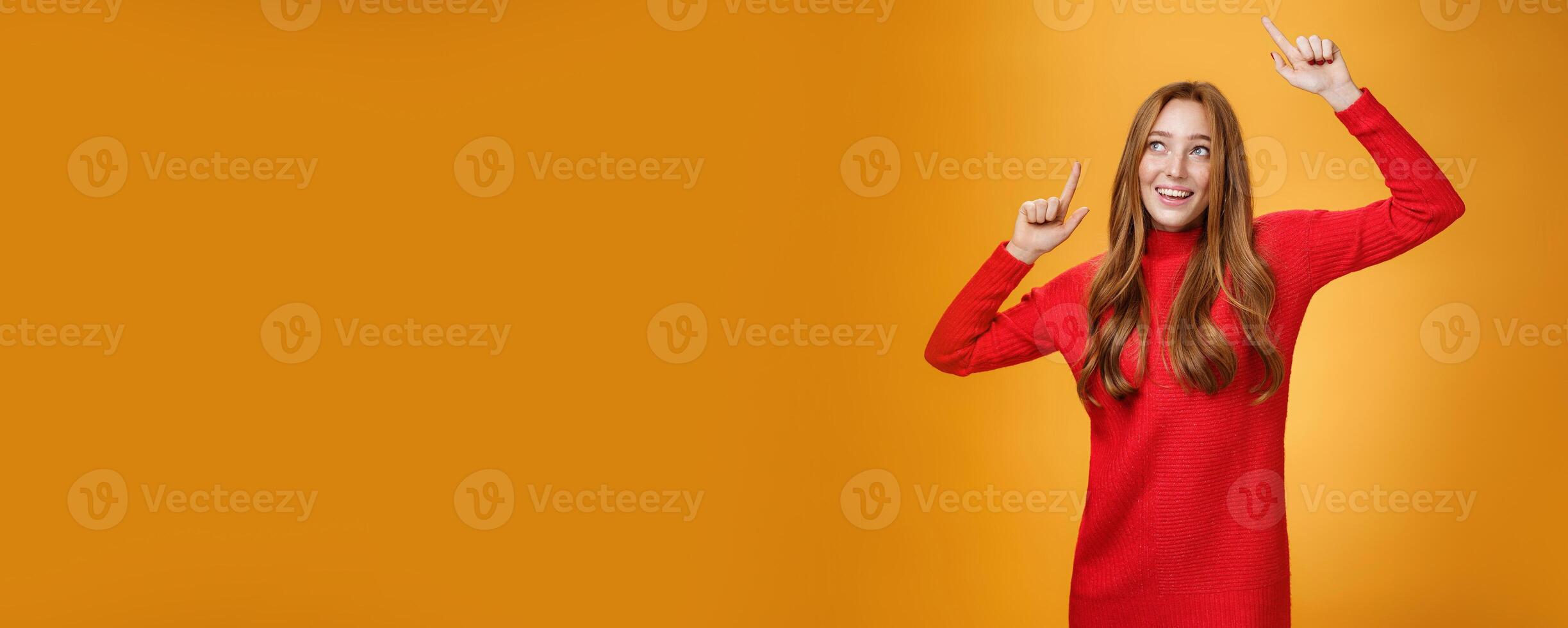 Charming and elegant attractive redhead female in red knitted dress raising hands up carefree as dancing having fun being pleased and happy gazing amused with broad grin at upper left corner photo
