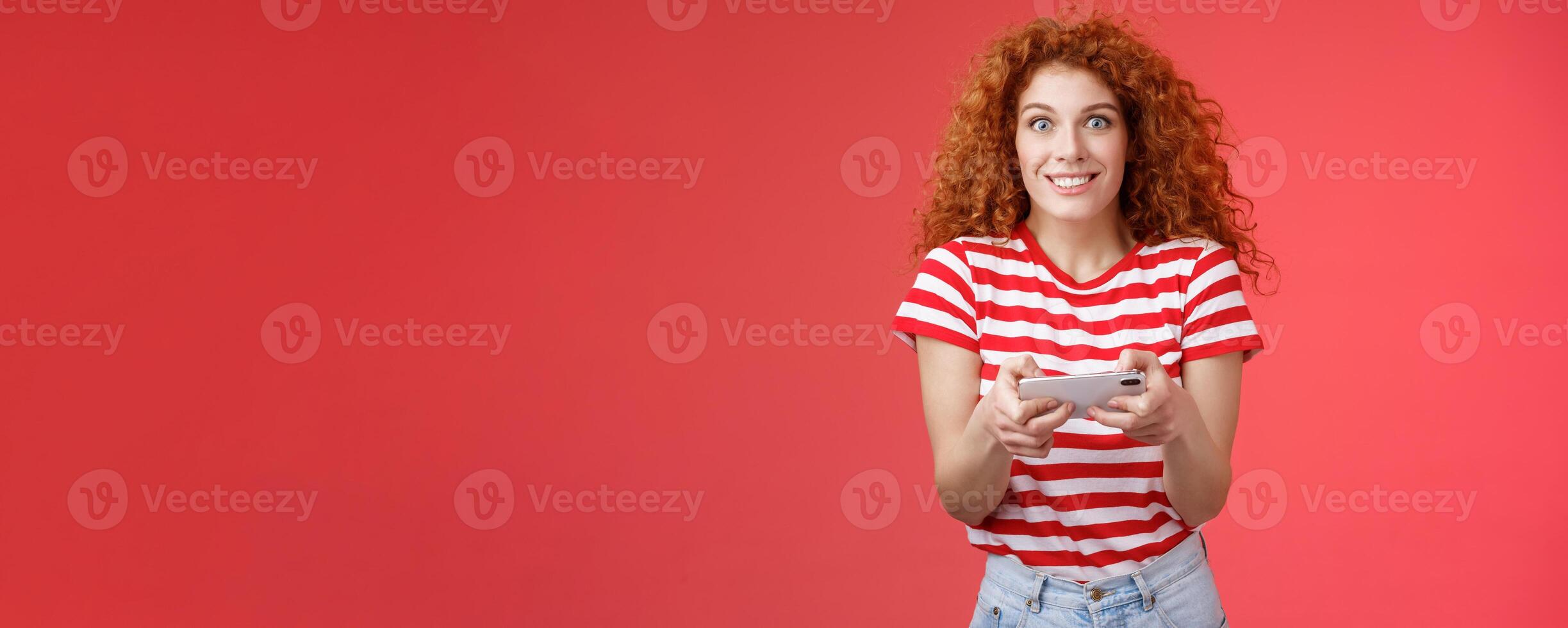 Excited happy cute redhead curly-haired girlfriend eager start learn how play boyfriend game look amused cheerful camera smiling joyful hold horizontal smartphone load app red background photo