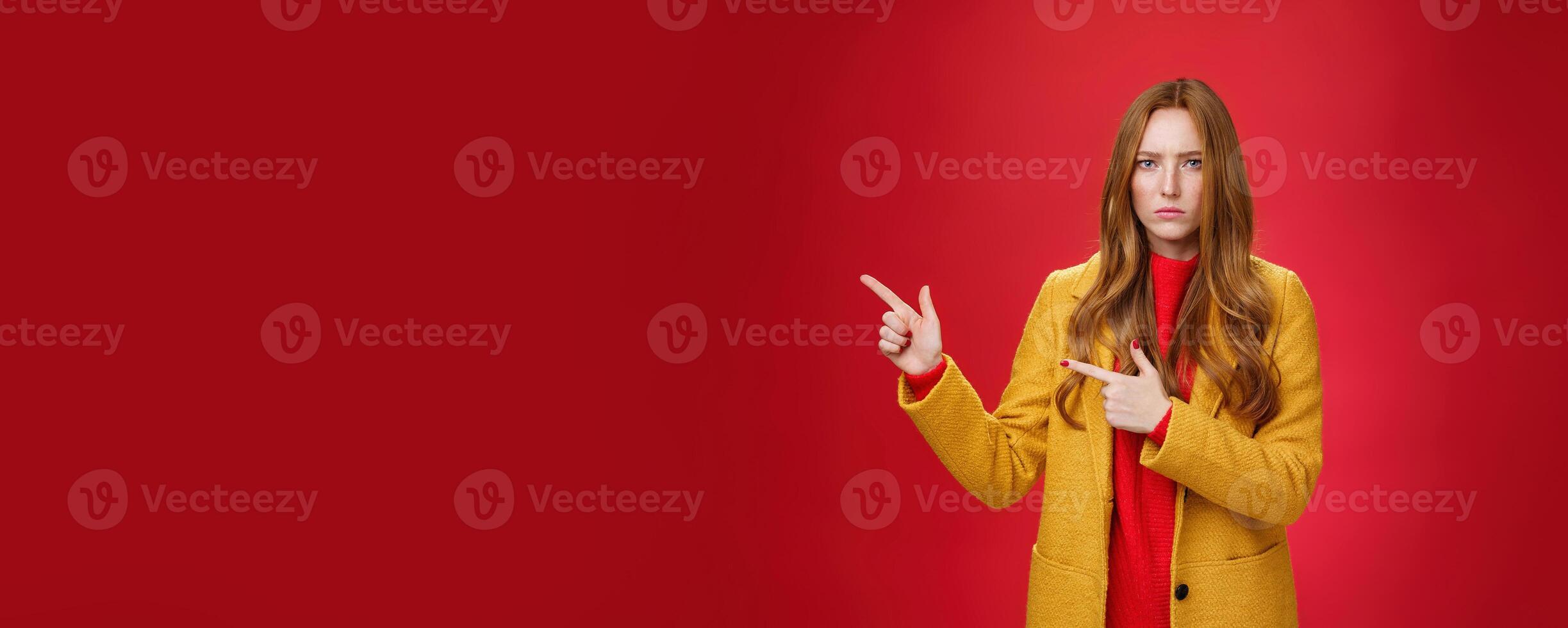 Displeased offended and gloomy girlfriend asking question, pouting and frowning from insult pointing at upper left corner disappointed and upset posing dissatisfied against red background photo