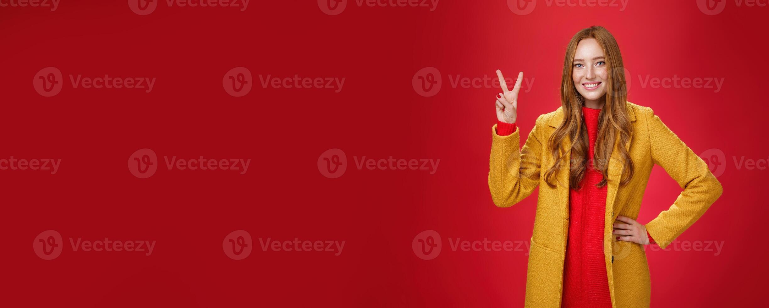 Portrait of happy playful and cute redhead woman with freckles in yellow coat holding hand on waist confident and self-assured showing peace or two sign delighted, beating weather conditions photo