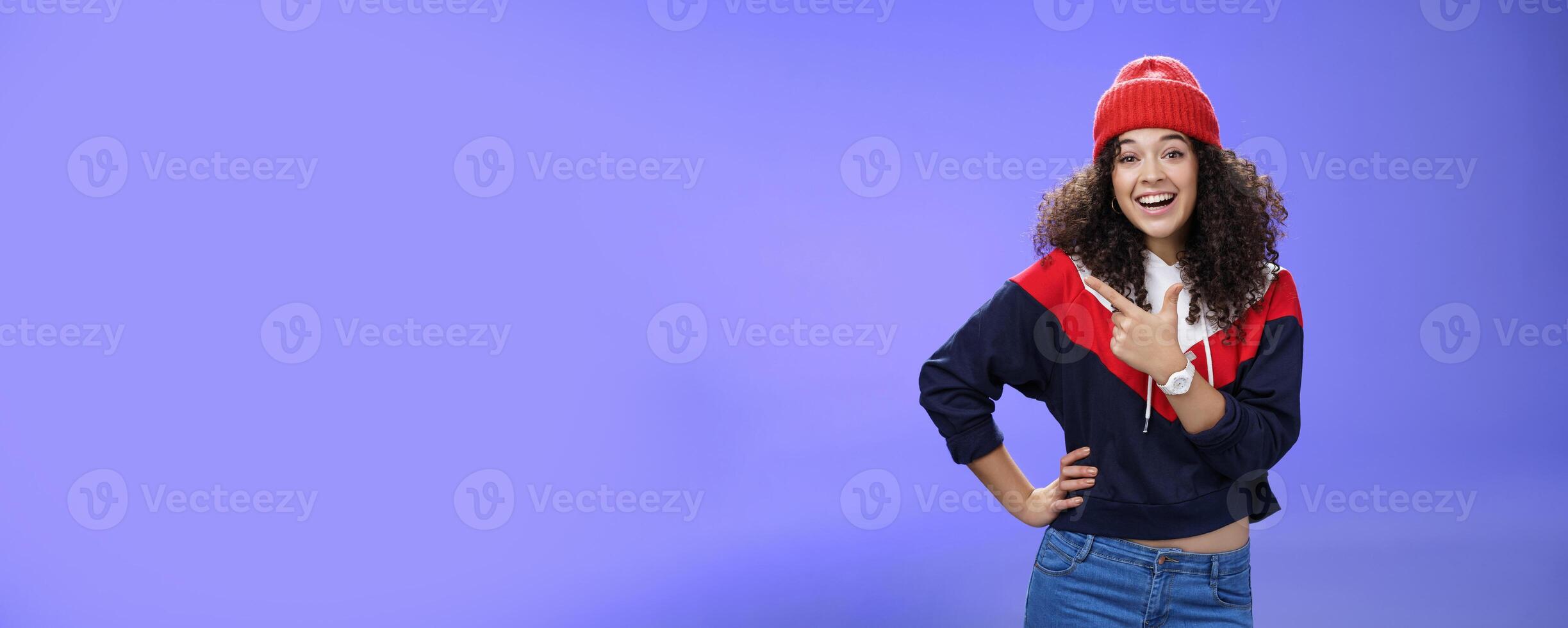 Stylish and sociable friendly-looking attractive woman with curly hair in warm beanie pointing at upper left corner laughing from joy and amazement having fun posing happily over blue background photo