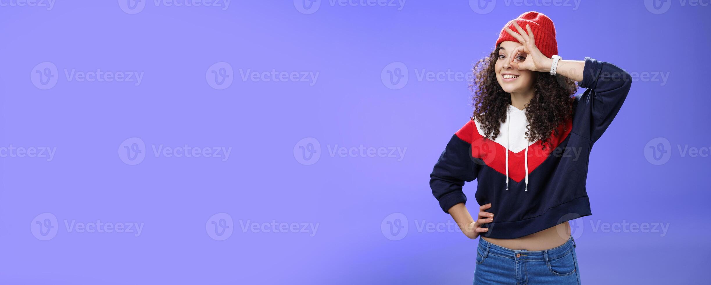 Okay girl agrees. Friendly-looking happy and charming young curly-haired girl in warm beanie showing ok gesture or circle over eye as peeking at camera and smiling broadly with joy over blue wall photo