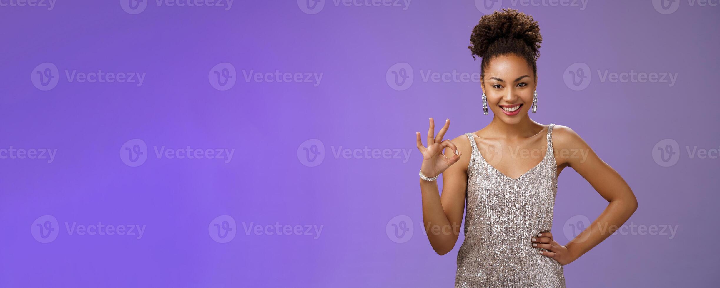 Girl shows everything under control. Sassy assertive african-american stylish woman in silver glittering dress show okay no problem gesture smiling self-assured guarantee all ok, blue background photo
