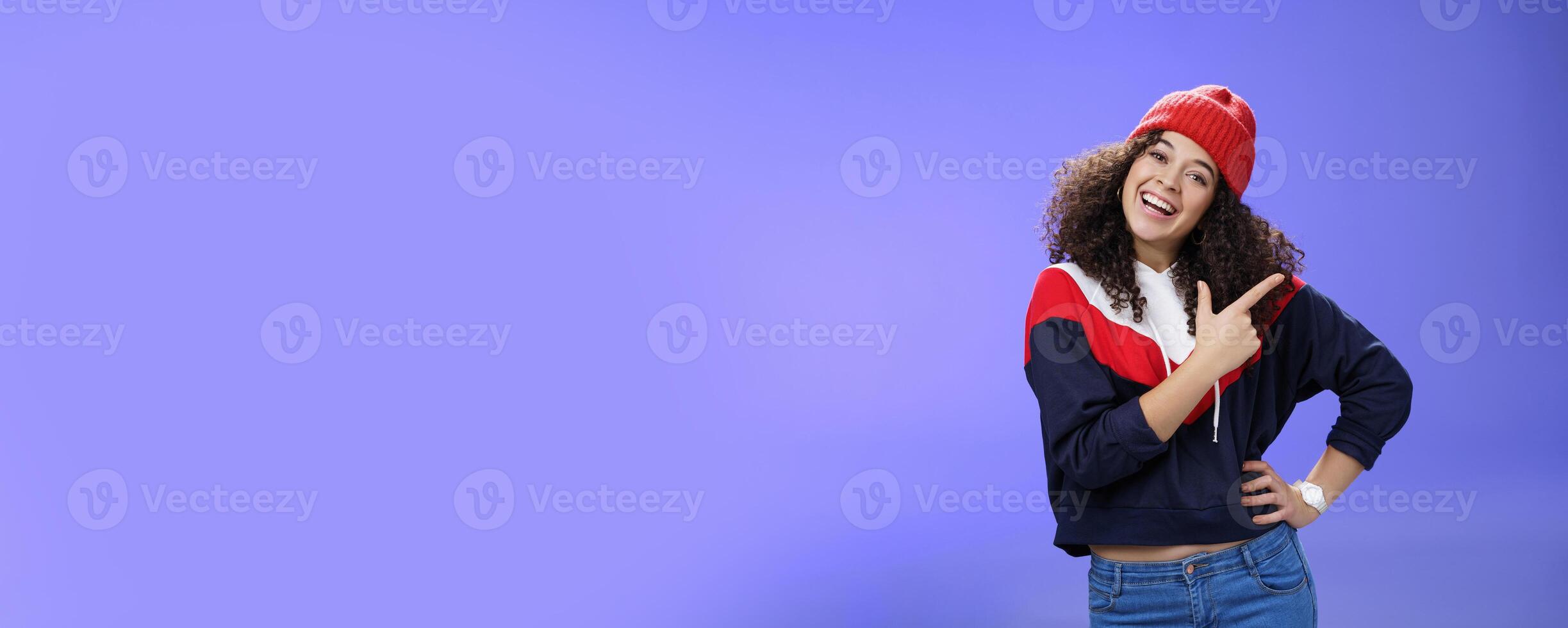 Studio shot of enthusiastic charming curly-haired woman inviting to play in snow with her pointing at upper right corner tilting head joyfully and smiling, holding hand on waist over blue wall photo