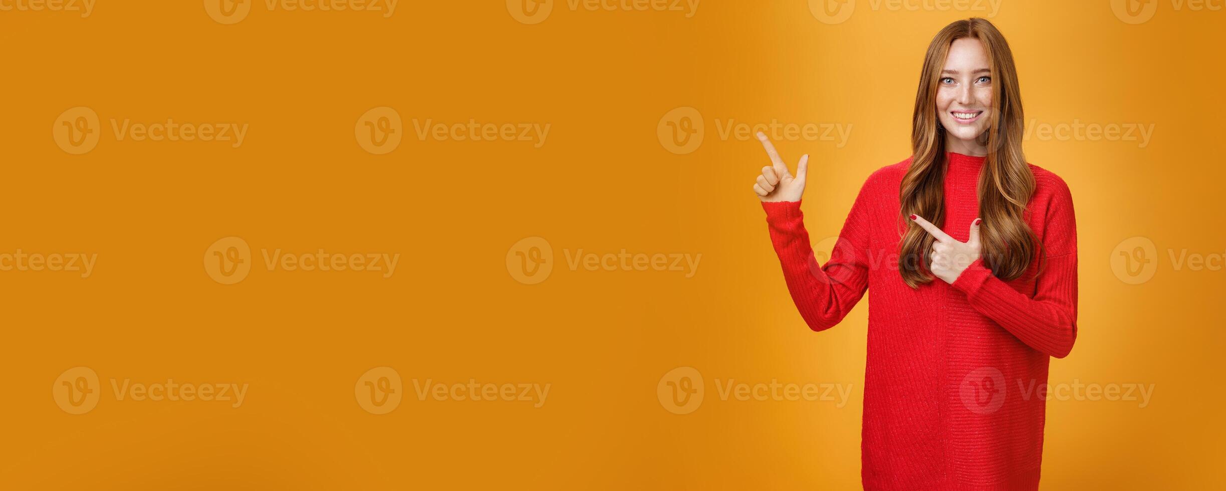 Assertive and helpful charming young female assitant in red knitted dress with ginger hair smiling friendly and pleasant as pointing at upper left corner, showing interesting thing over orange wall photo