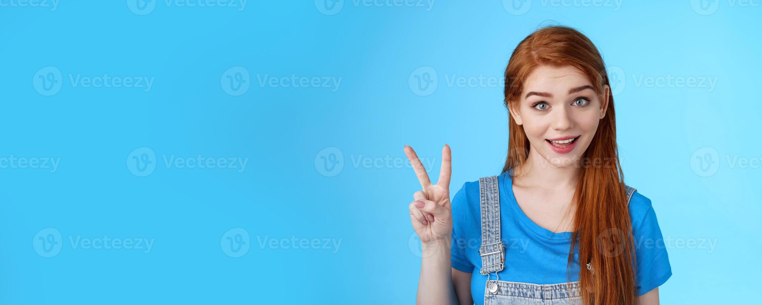 Cheerful attractive redhead caucasian girl smiling joyfully talking, order two peaces, show second number, peace victory sign, stand carefree, upbeat emotions, stand blue background photo