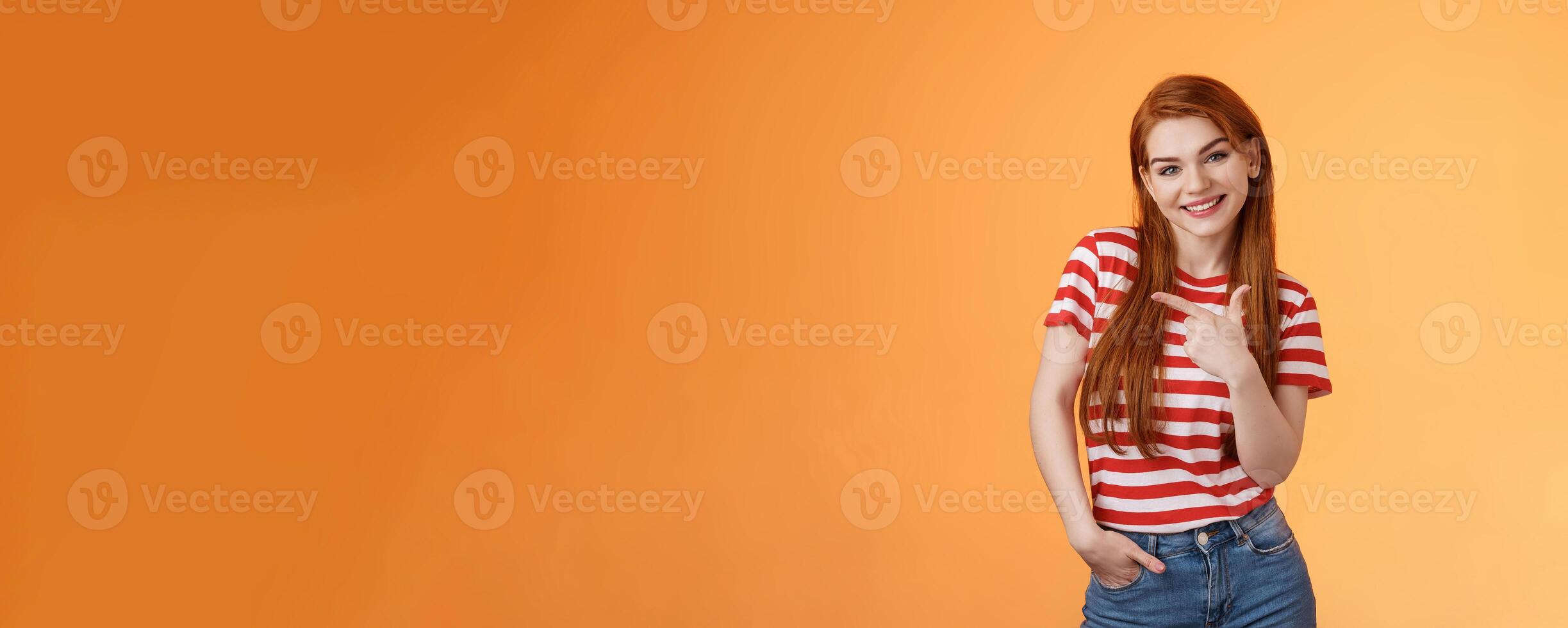 Tender cute feminine redhead lovely girl, tilt head cheerfully smiling, pointing left, showing way, give advice what purchase, advertising good product, introduce shop, orange background photo