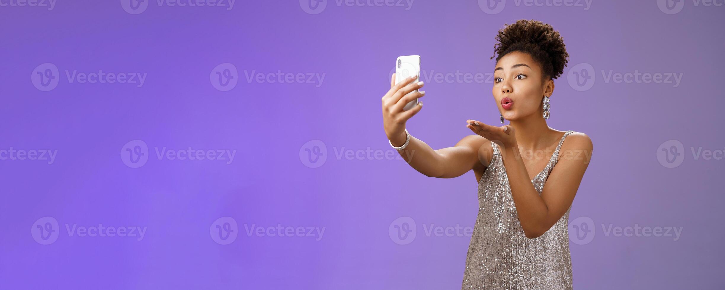 Charming elegant young female student partying enjoying prom recording smartphone look phone display send air kiss blowing mwah taking selfie hold gadget, standing blue background photo