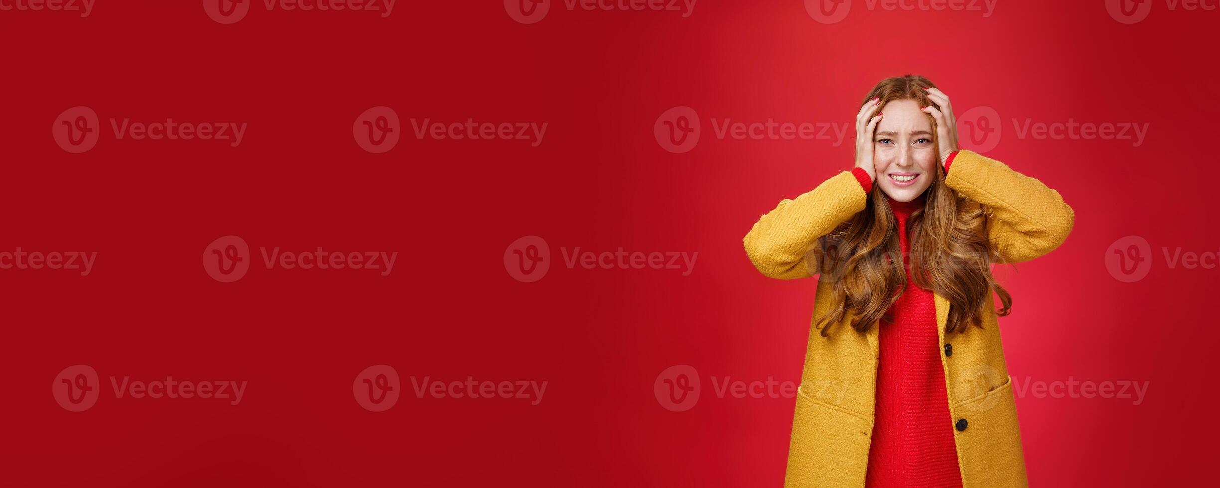 Portrait of redhead woman in panic holding hands pressed to head clenching teeth and frowning puzzled and troubled, standing anxious and concerned freaking out over red background photo