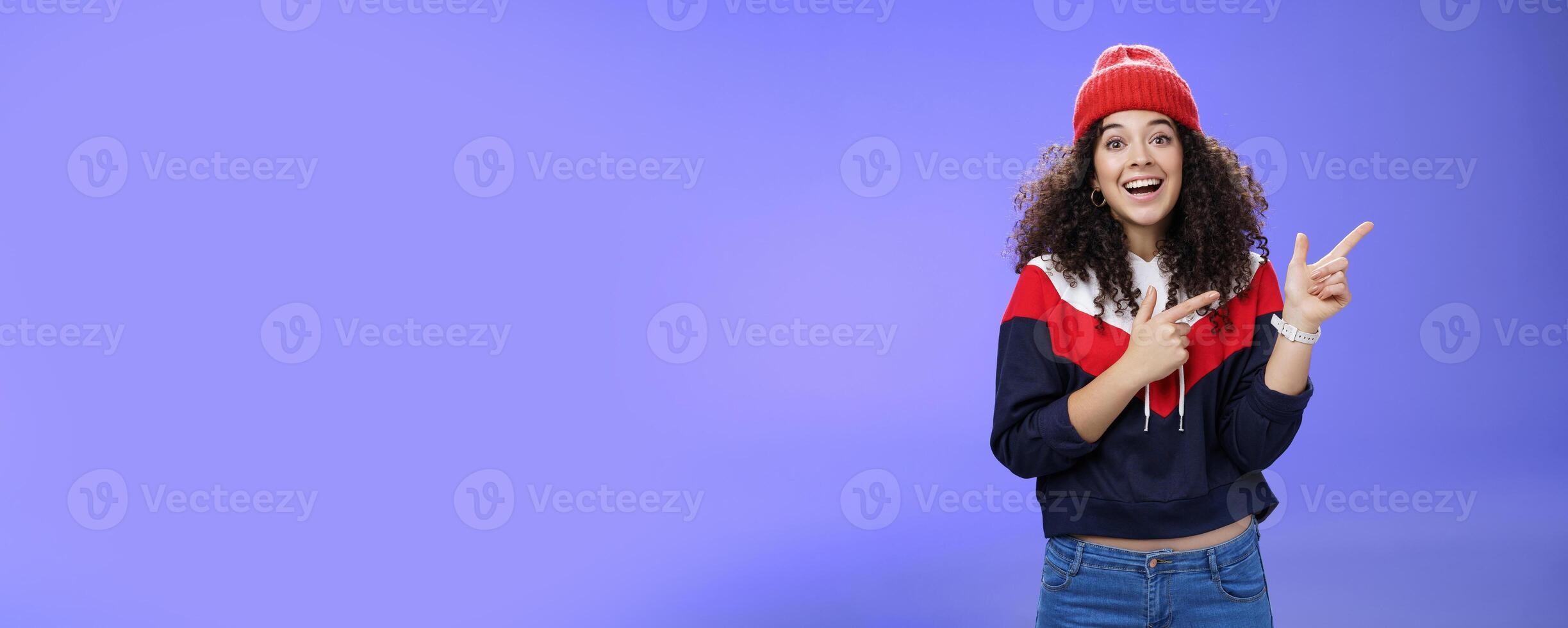Enthusiastic friendly-looking attractive young 20s woman with curly hair in warm beanie and sweatshirt smiling open mouth amazed and delighted as pointing at upper right corner over blue background photo