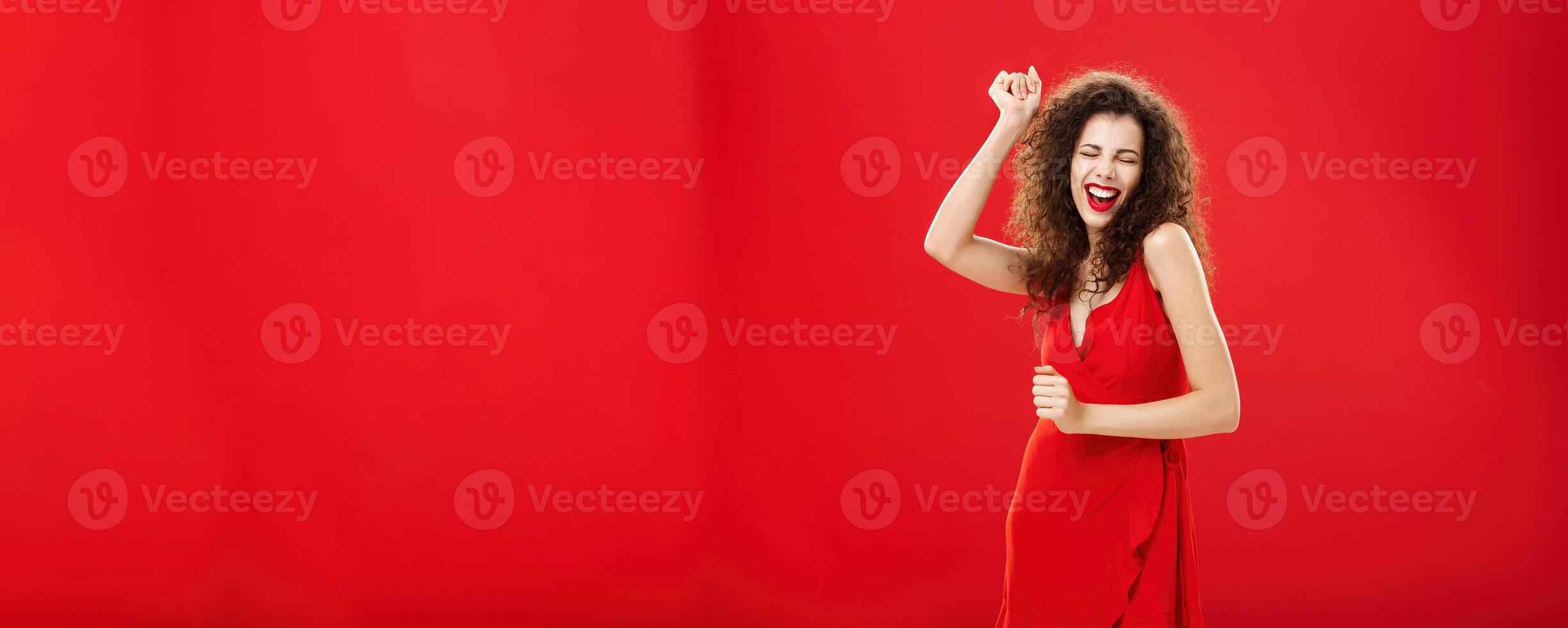 Rich woman forgetting all troubled on dance floor. Charming elegant young european female model in stylish evening dress moving to rhythm of music rejoicing raising hand, singing posing over red wall photo