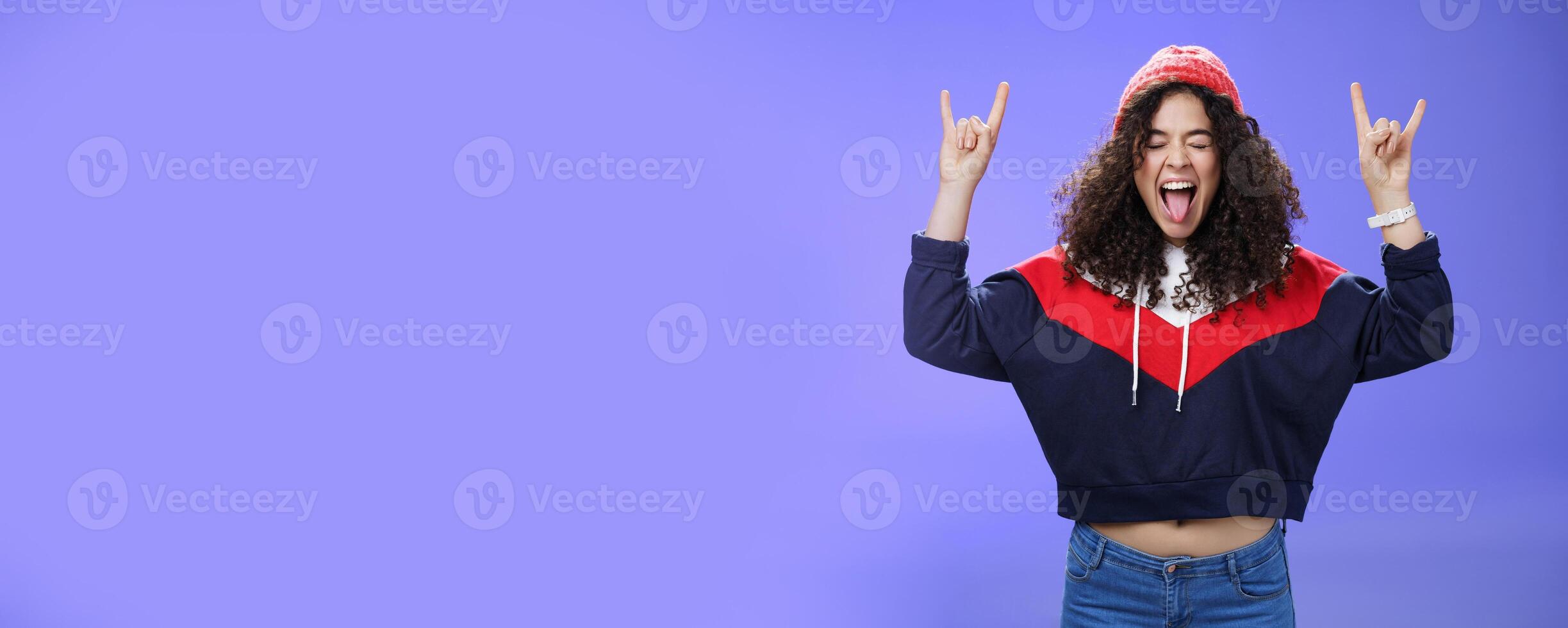 Waist-up shot of rebellious and carefree woman having fun feeling excited and awesome at party close eyes sticking out tongue and showing rock-n-roll signs, wearing beanie and warm clothes photo