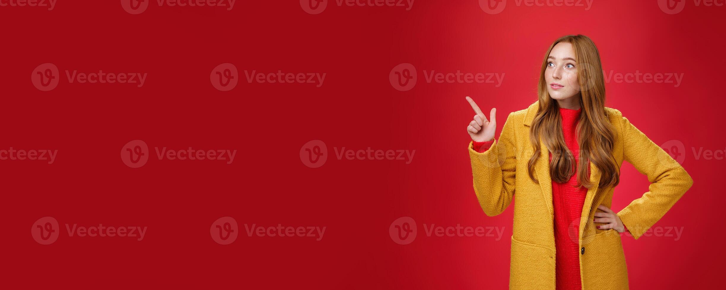 Wondered curious good-looking redhead woman in coat stopping on way looking and pointing at upper left corner curious and interested, observing surprising promotion over red background photo