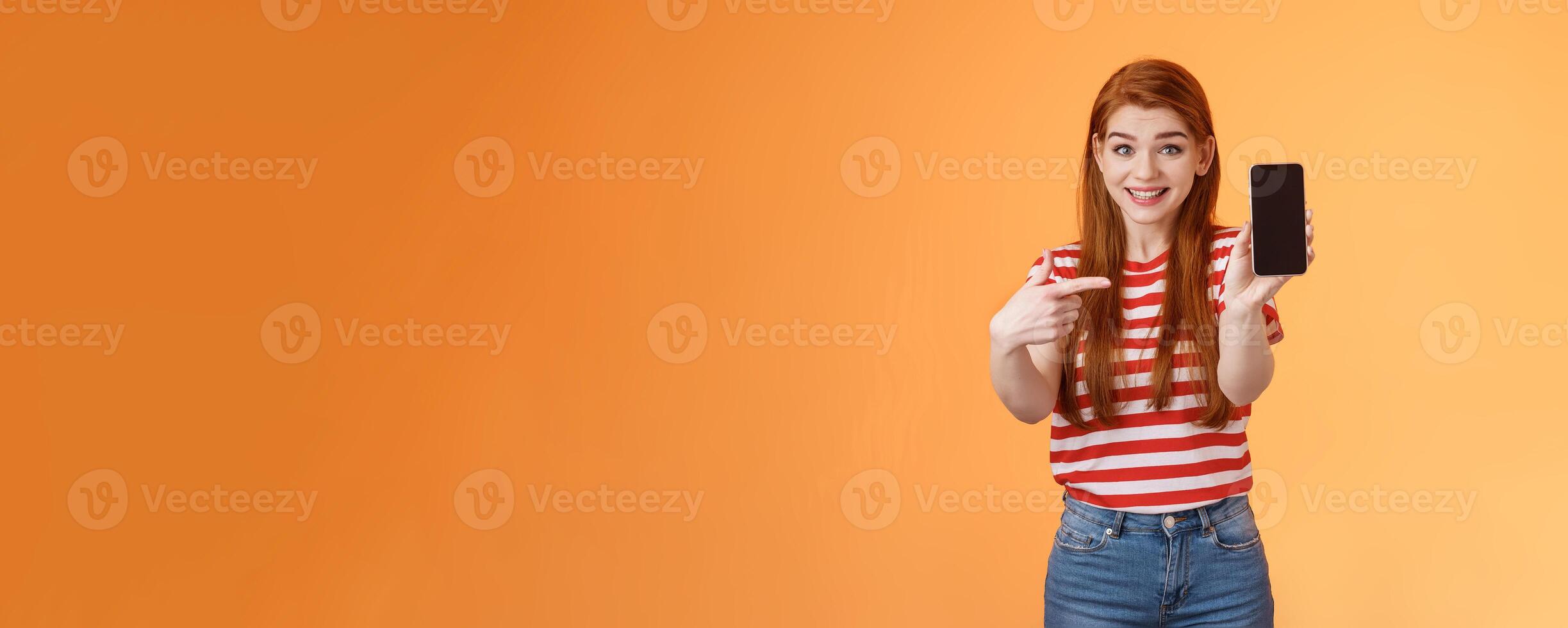 Impressed excited happy redhead woman smiling broadly, hold smartphone, pointing cellphone screen, thrilled amazed introduce fascinating application, stand orange background rejoice photo
