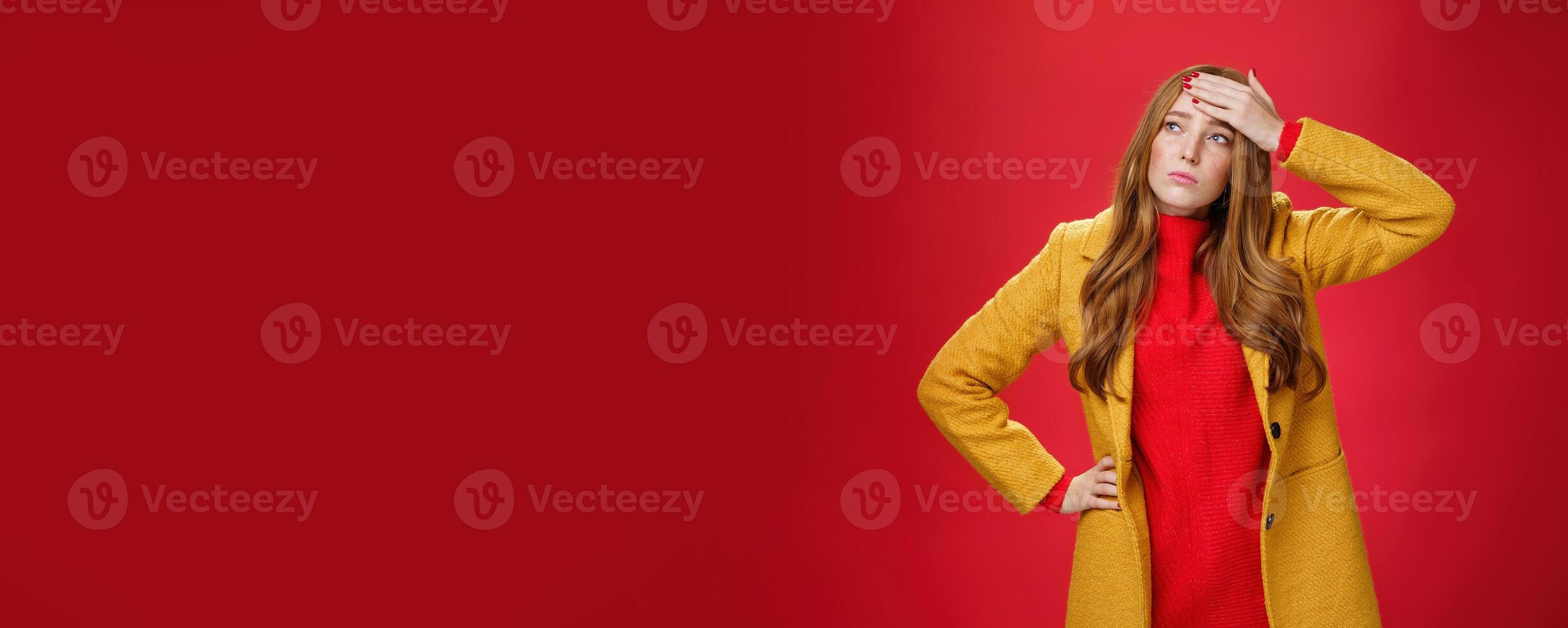 Girl having feaver touching forehead with palm looking displeased and bothered left as thinking she got cold standing in yellow coat, realising she sick or ill over red background, feeling tired photo