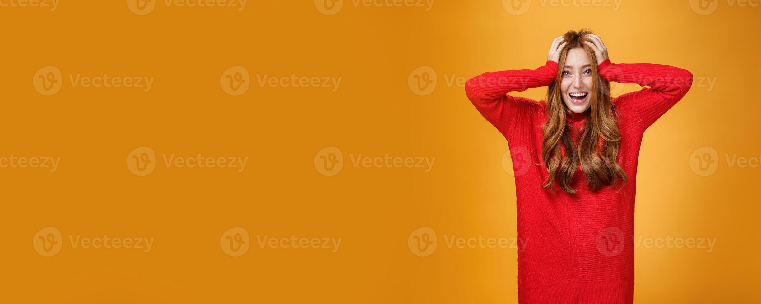 Excited and overwhelmed charming redhead woman in knitted warm red dress yelling from thrill and happiness holding hands on hair smiling broadly impressed with unexpected luck over orange wall photo
