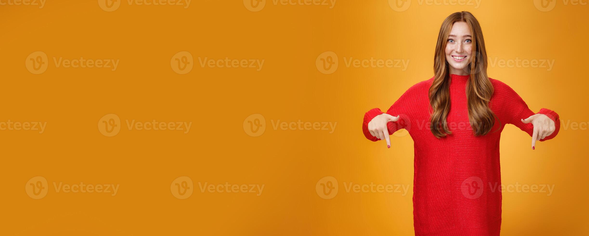 Gentle and tender cute ginger girl with freckles and blue eyes feeling touched and pleased, pointing at heartwarming promotion, showing direction down and smiling against orange background photo