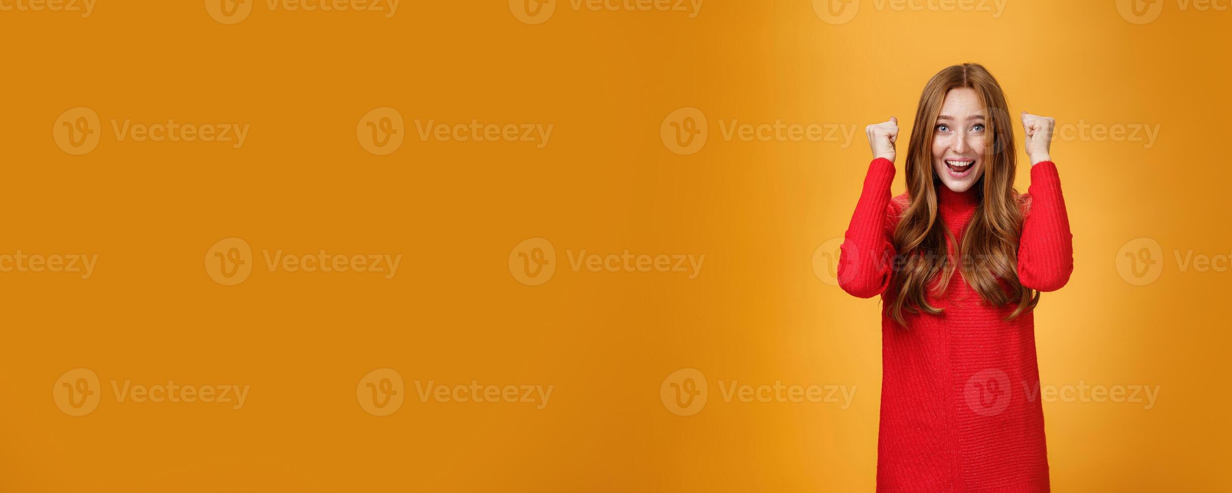 Happy delighted and cheerful ginger girl with freckles and blue eyes triumphing yelling yes with broad smile clenching raised fists in joy and celebration of success against orange background photo
