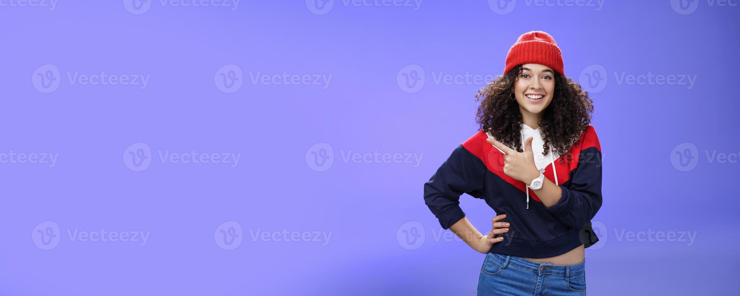 Studio shot of happy charming stylish city girl in beanie and warm sweatshirt ready rock winter up pointing at upper left corner and smiling delighted showing cool place over blue wall photo