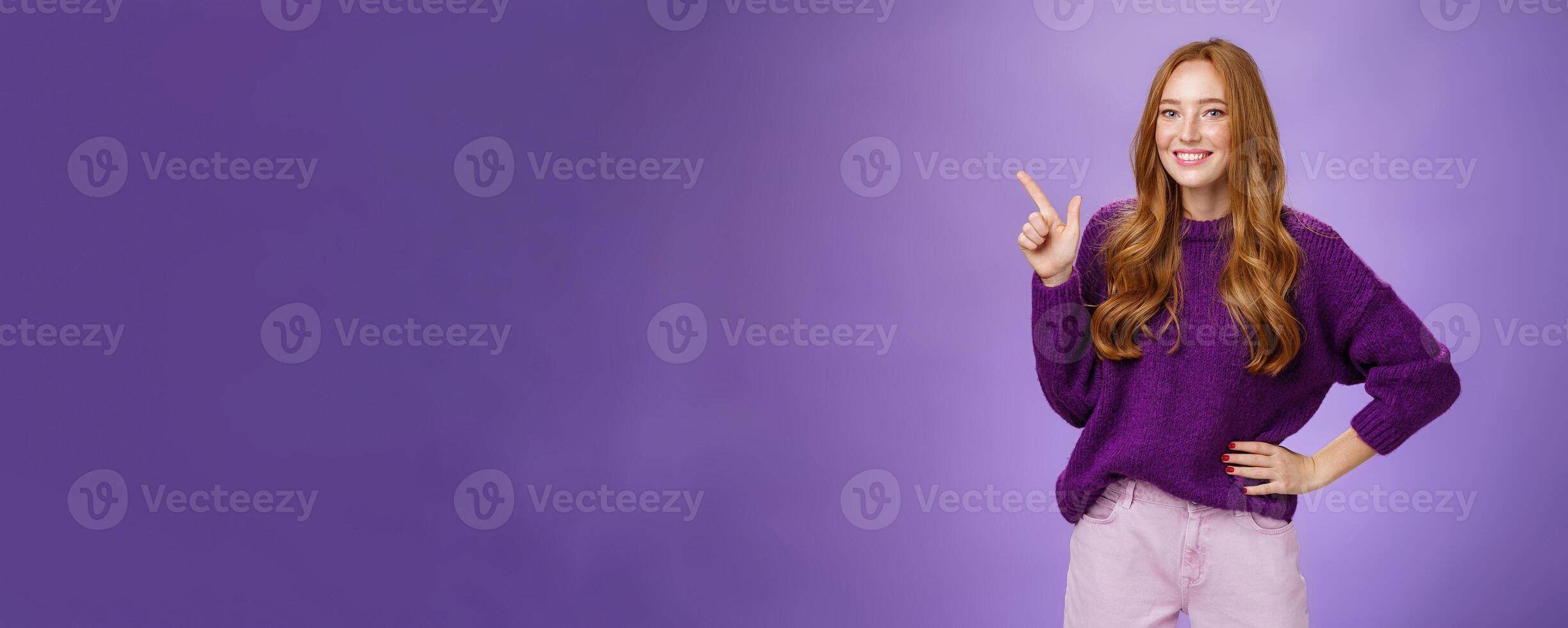 Hey click here. Portrait of friendly-looking charming and cute redhead female in purple sweater pointing at upper left corner with index finger and smiling, showing copy space, recommending, offering photo