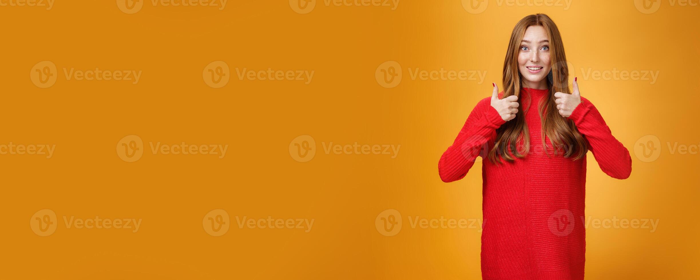 Excited and supportive redhead girlfriend asking opinion about new warm dress showing thumbs up and raising eyebrows questioned as waiting for opinion over orange background photo