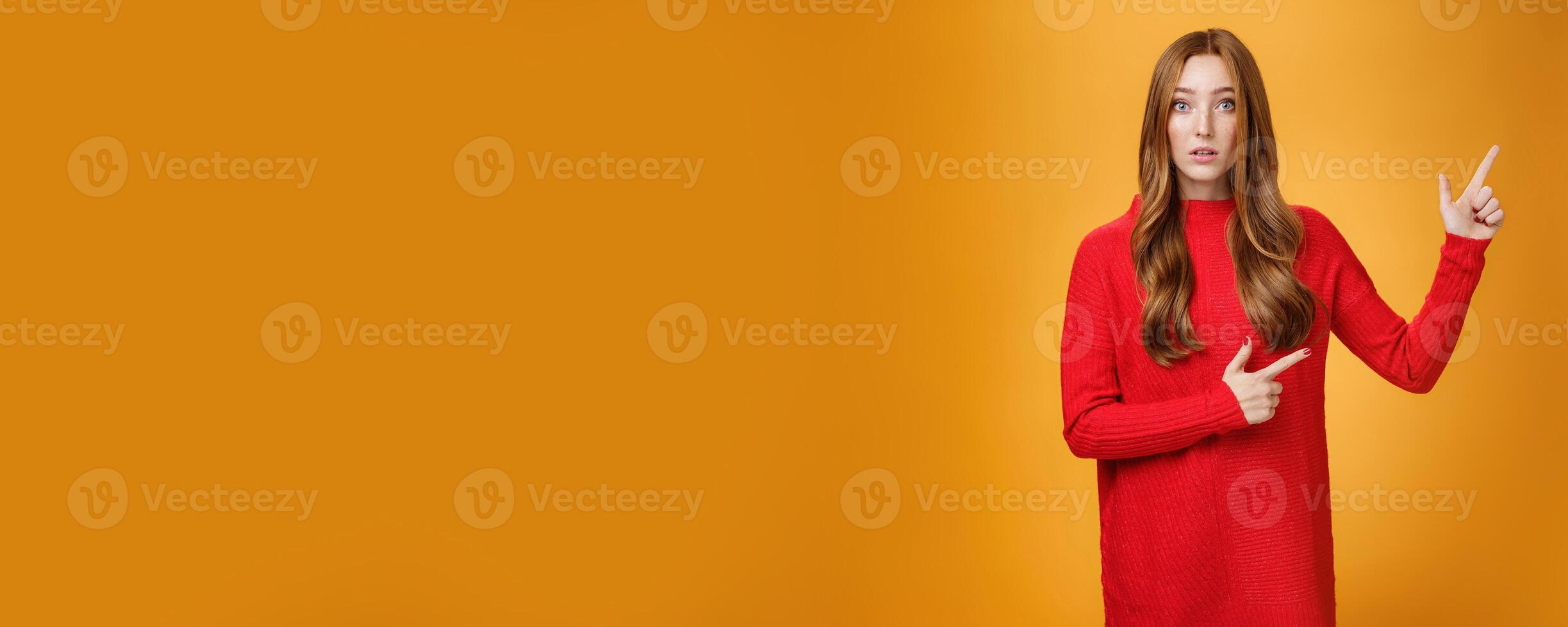 Portrait of elegant and stylish redhead 20s woman with freckles and long hair in knitted red dress pointing at upper right corner looking questioned and impressed, speechless over orange background photo