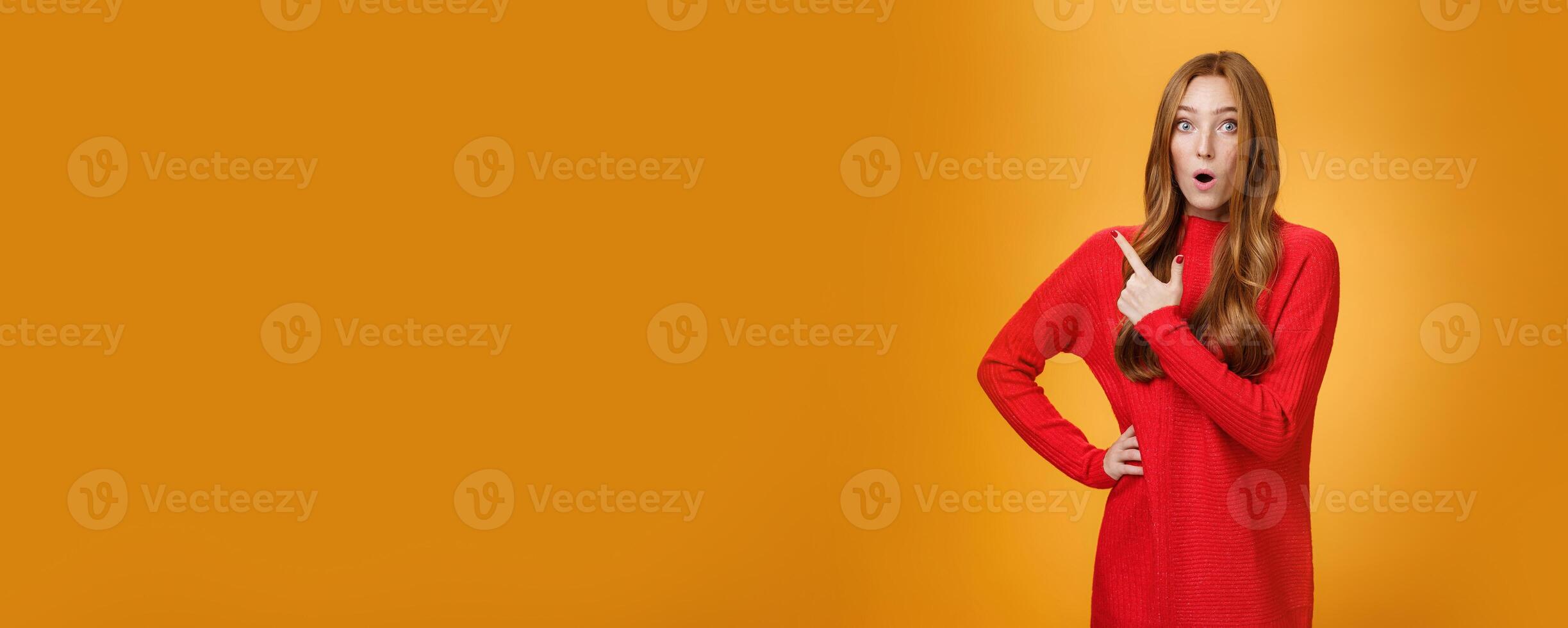 Impressed redhead girl reacting to awesome unbeliavable promotion pointing behind or at upper left corner folding lips staring at camera astonished and surprised over orange background photo