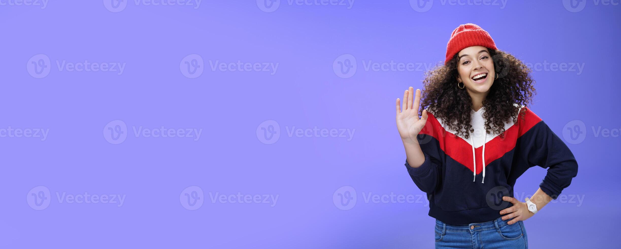 Hey nice to meet you. Charming female ski couch in cute red beanie with curly hair waving hello with palm and smiling broadly greeting newbies as teaching winter sports over blue background photo