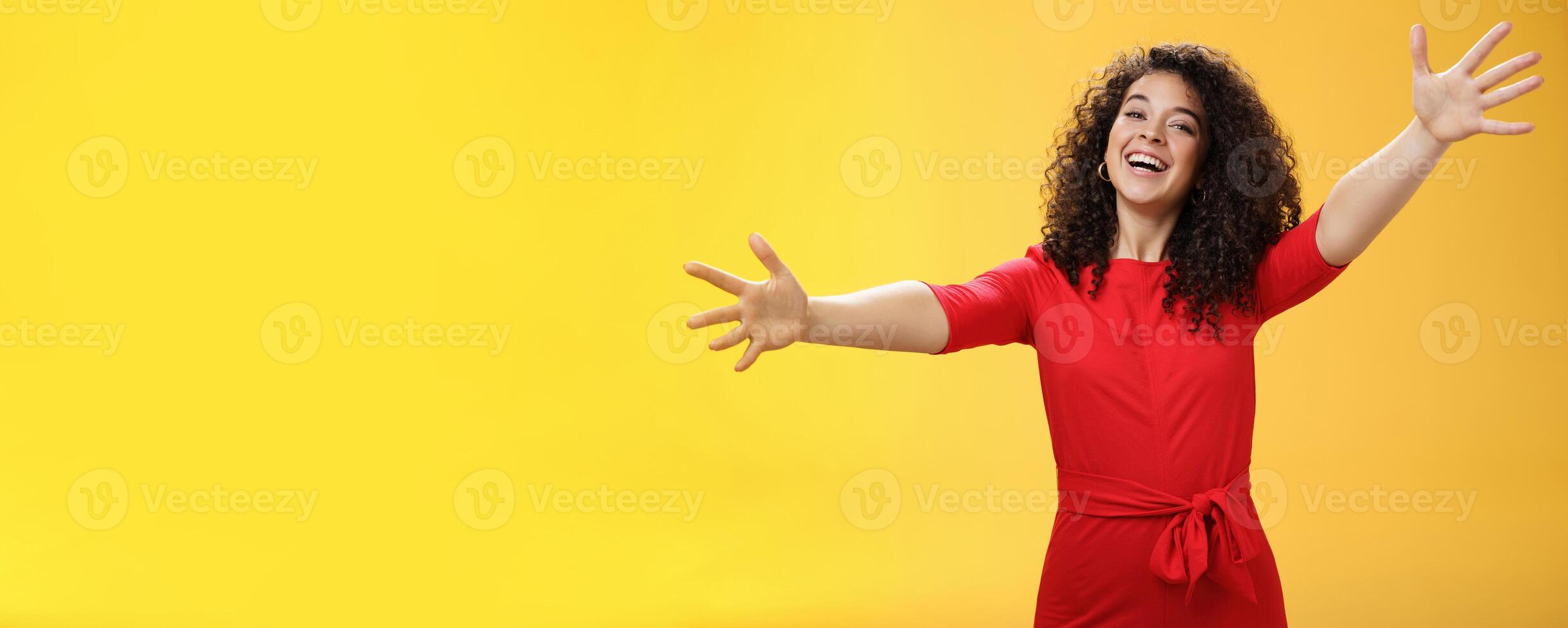 Come into my arms. Portrait of friendly and loving, caring charming woman with curly hair in red casual dress spread hands as wanting give hug smiling broadly at camera giving warm welcome or cuddle photo