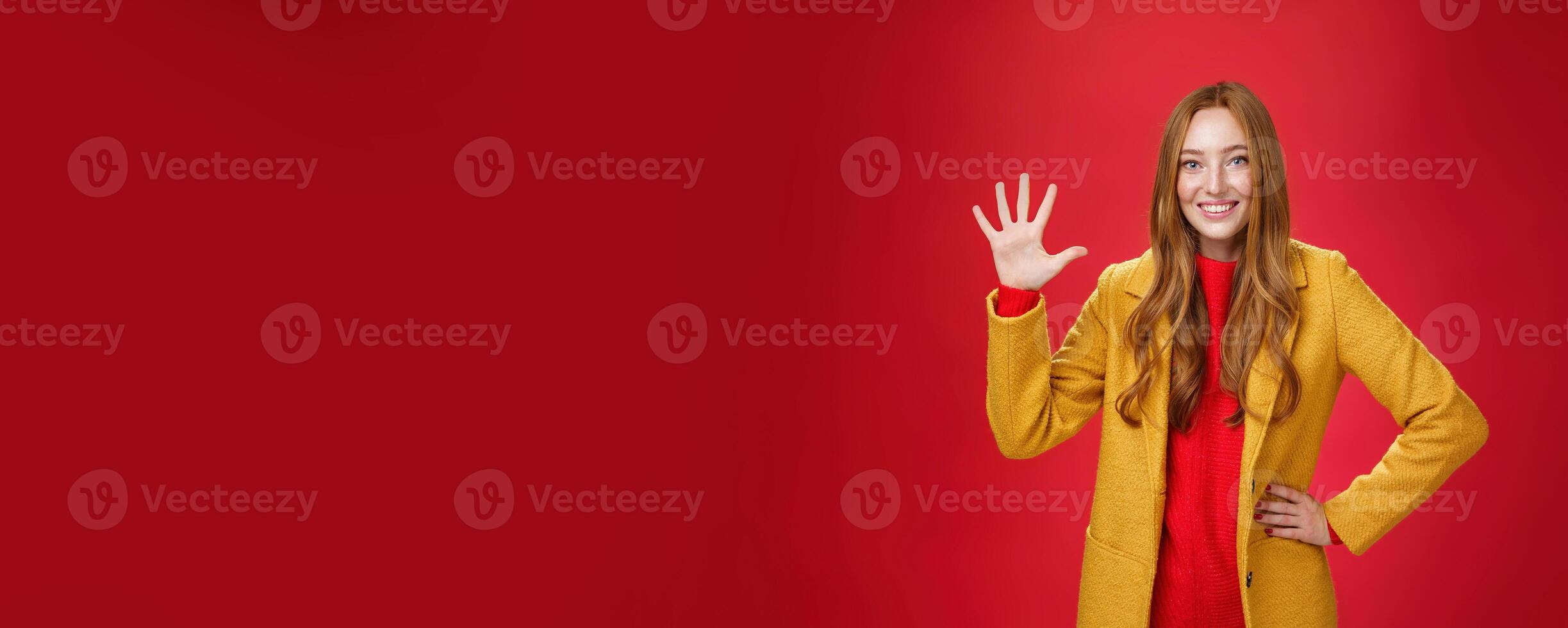 Girl giving five stars to awesome internet store she bought outfit for autumn weather showing raised palm smiling broadly as standing in stylish warm yellow coat over red background photo