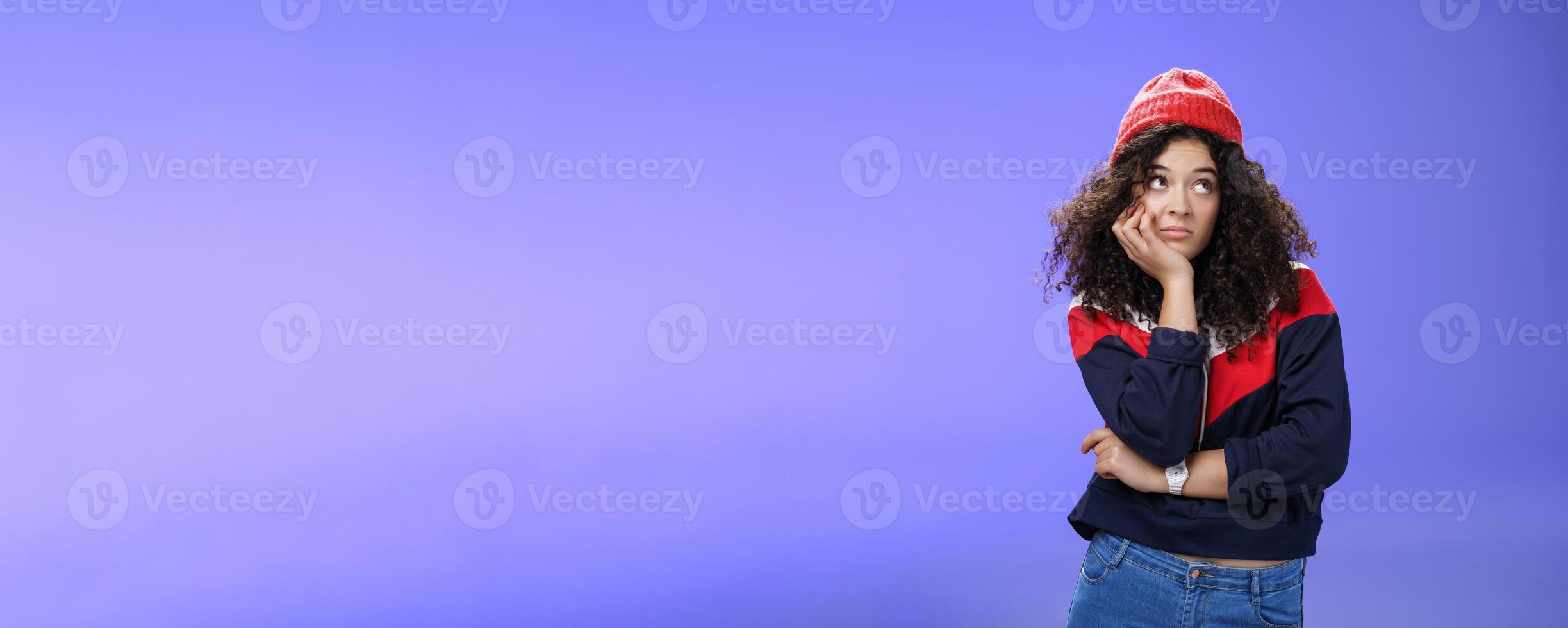 Portrait of frustrated and bored lonely girl with curly hair in winter hat holding hand across chest leaning head on palm, looking silly and gloomy at upper right corner, thinking feeling boredom photo