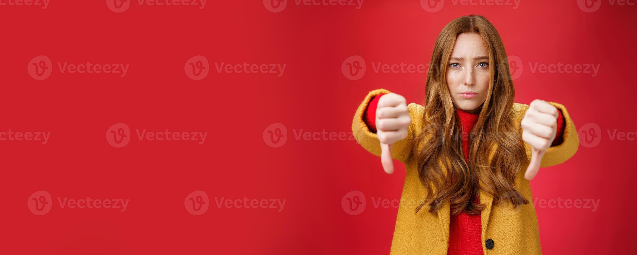 Portrait of insecure and unconfident cute redhead female reacting to uncool thing showing thumbs down with worried upset expression from dislike and disappointment over red background photo