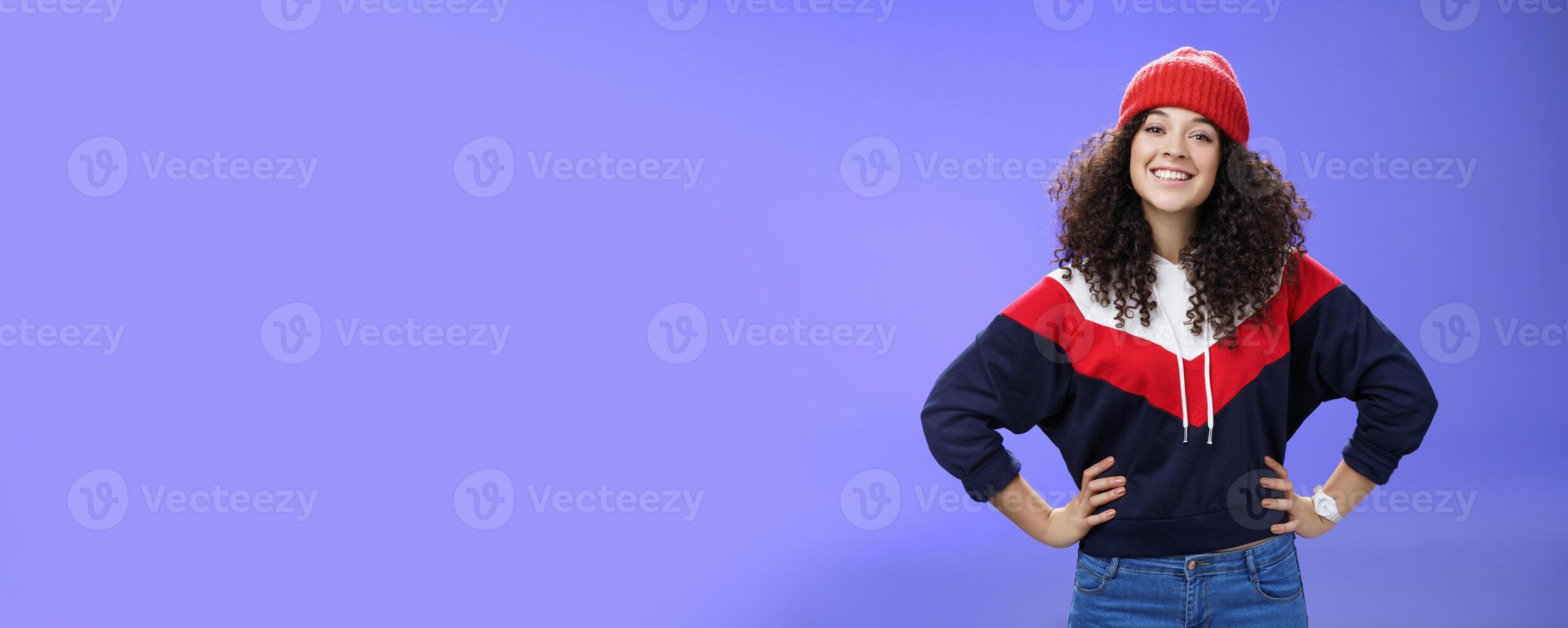Confident ad energized cute european woman with curly hairstyle in warm beanie and stylish sweatshirt holding hands on waist and smiling friendly at camera as ready to go out and have fun outside photo