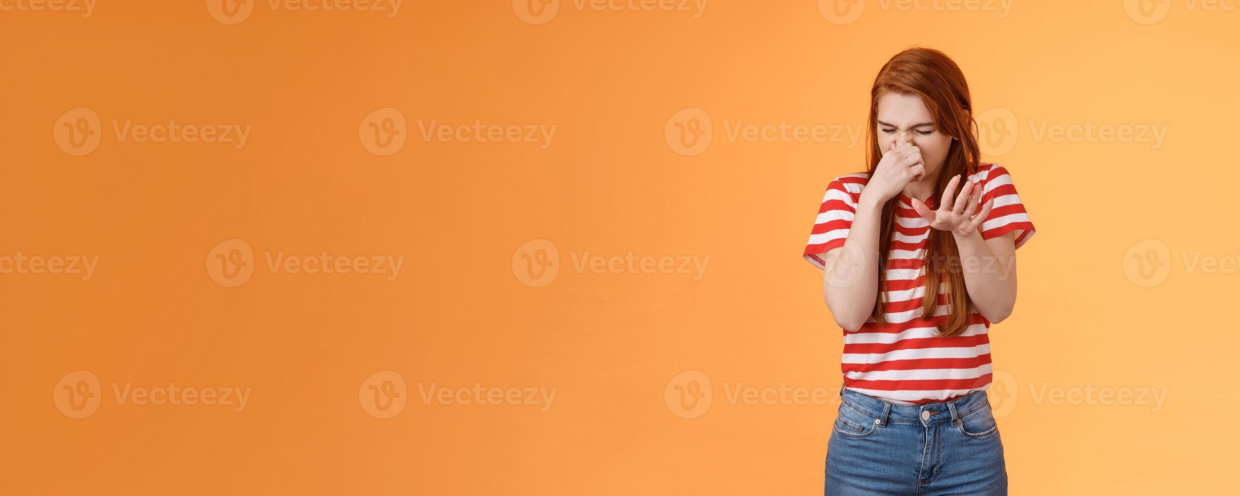 Gosh it stinks. Reluctant disgusted redhead woman raise hand block dislike gesture shut nose, cringe from aversion dislike, puke from reek bad smell, stand orange background disappointed photo
