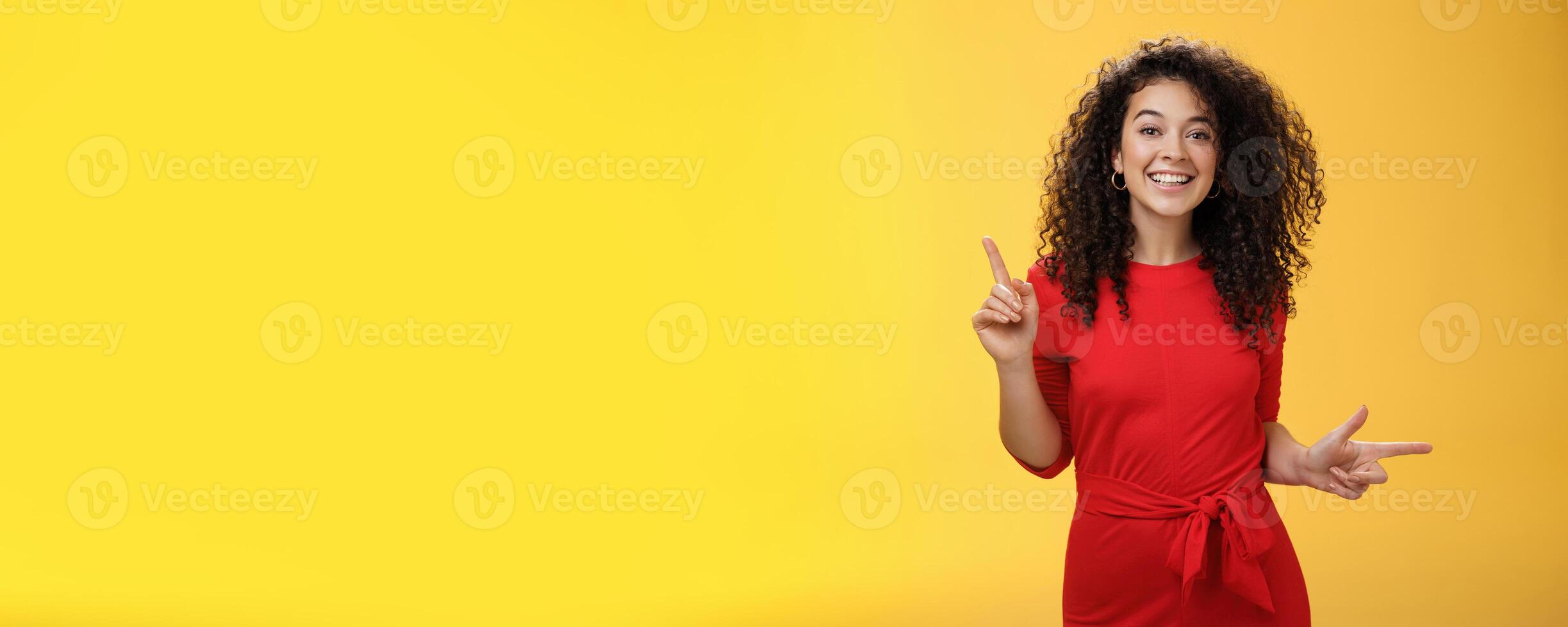 Charismatic cute and happy young curly-haired european female in red dress dancing and pointing up and right, showing sideways as presenting choices picking, making decision and smiling at camera photo