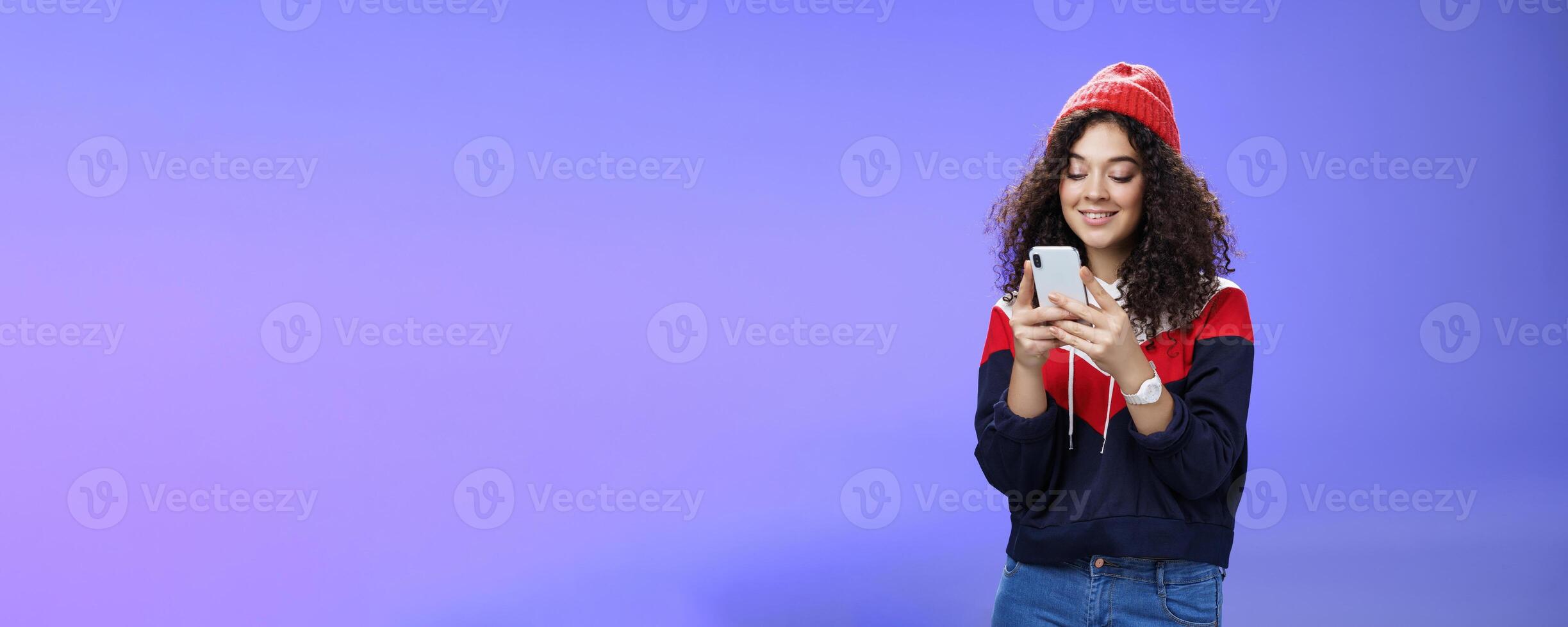 Woman searching in internet funny memes, holding mobile phone in hands smiling joyfully and cute at smartphone screen, typing message or browsing internet over blue background, wearing warm winter hat photo